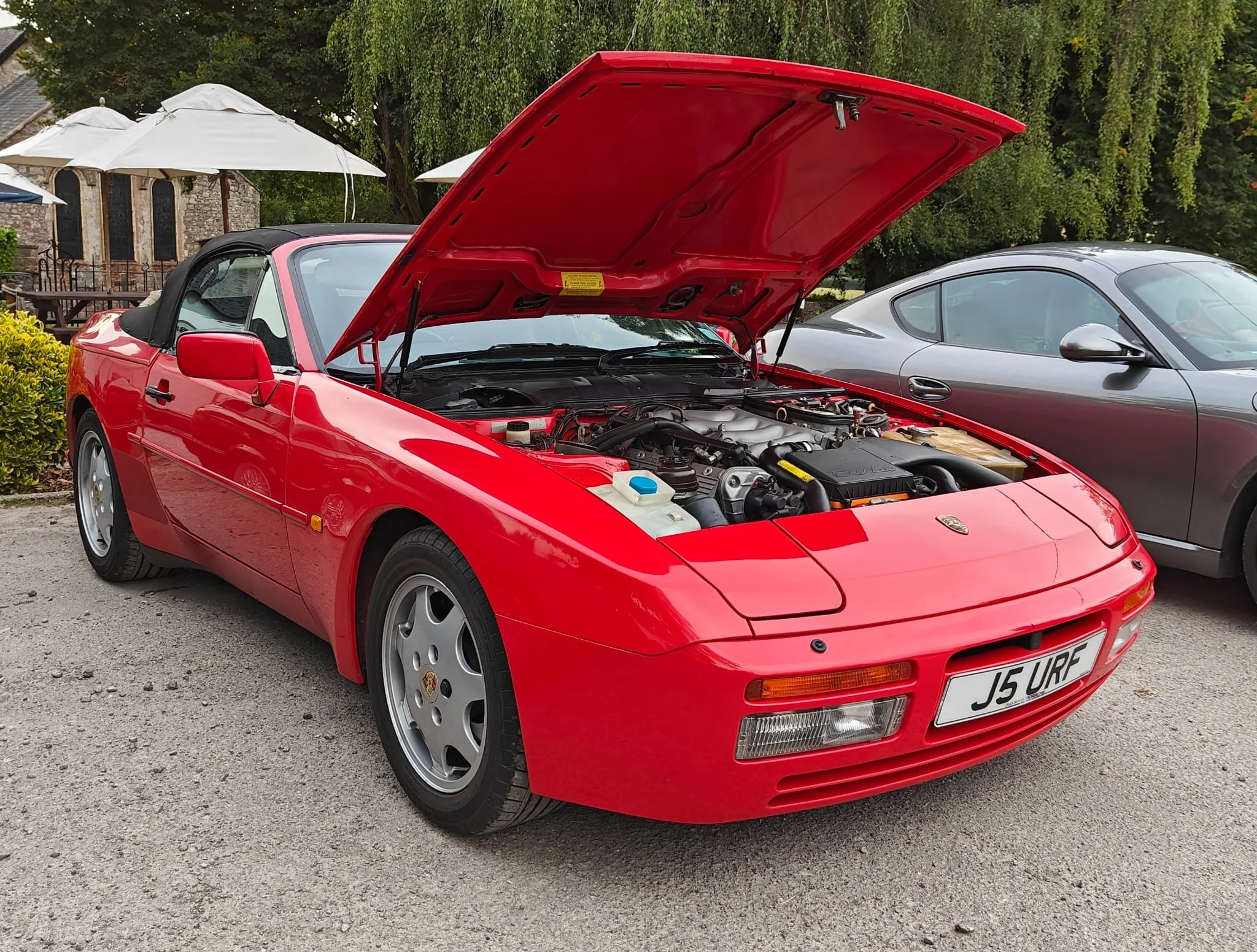 A red Porsche sports car with its hood open, exposing the engine, parked on a gravel surface next to a gray sports car in a scenic outdoor setting with trees and umbrellas.