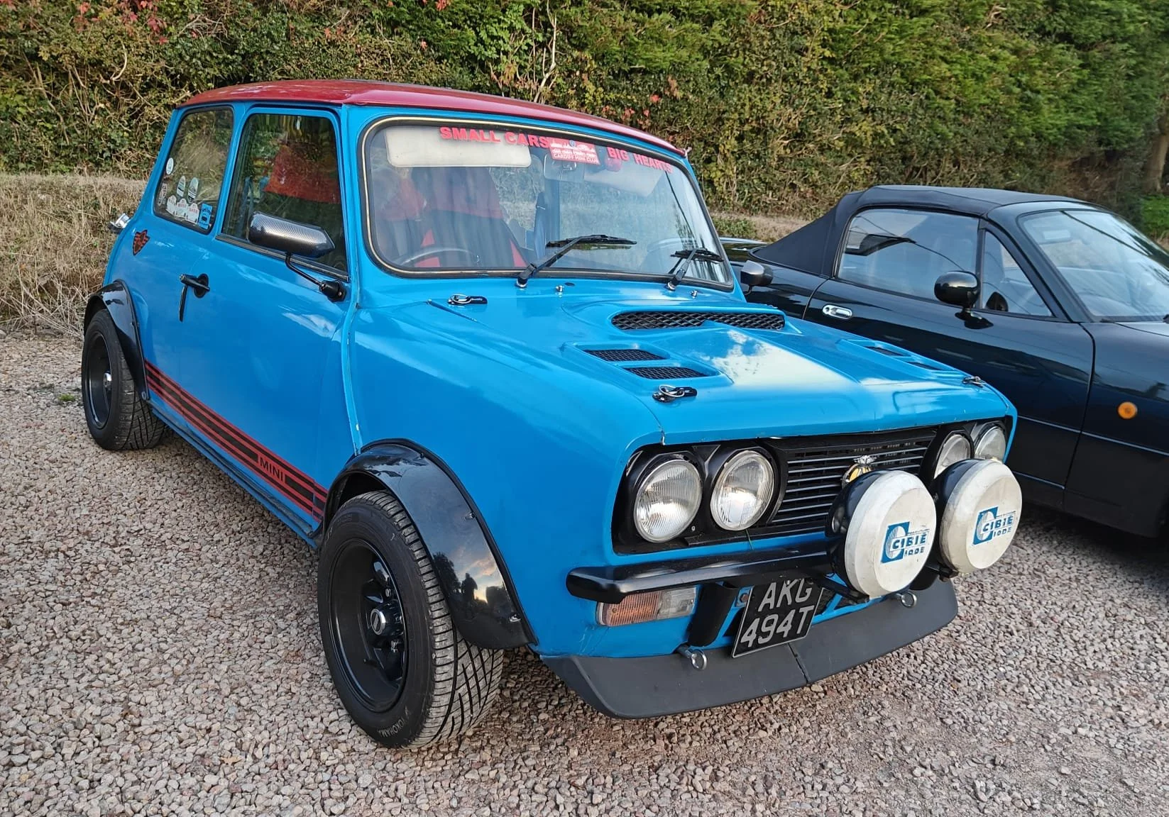 A vintage blue Mini Cooper with rally lights, black fender flares, and decals parked outdoors on a gravel surface next to a black convertible car.