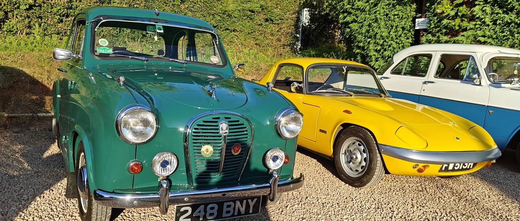 Three vintage cars parked on a gravel surface with green foliage in the background. A green classic car on the left, a yellow sports car in the middle, and a partially visible white and blue vehicle on the right.