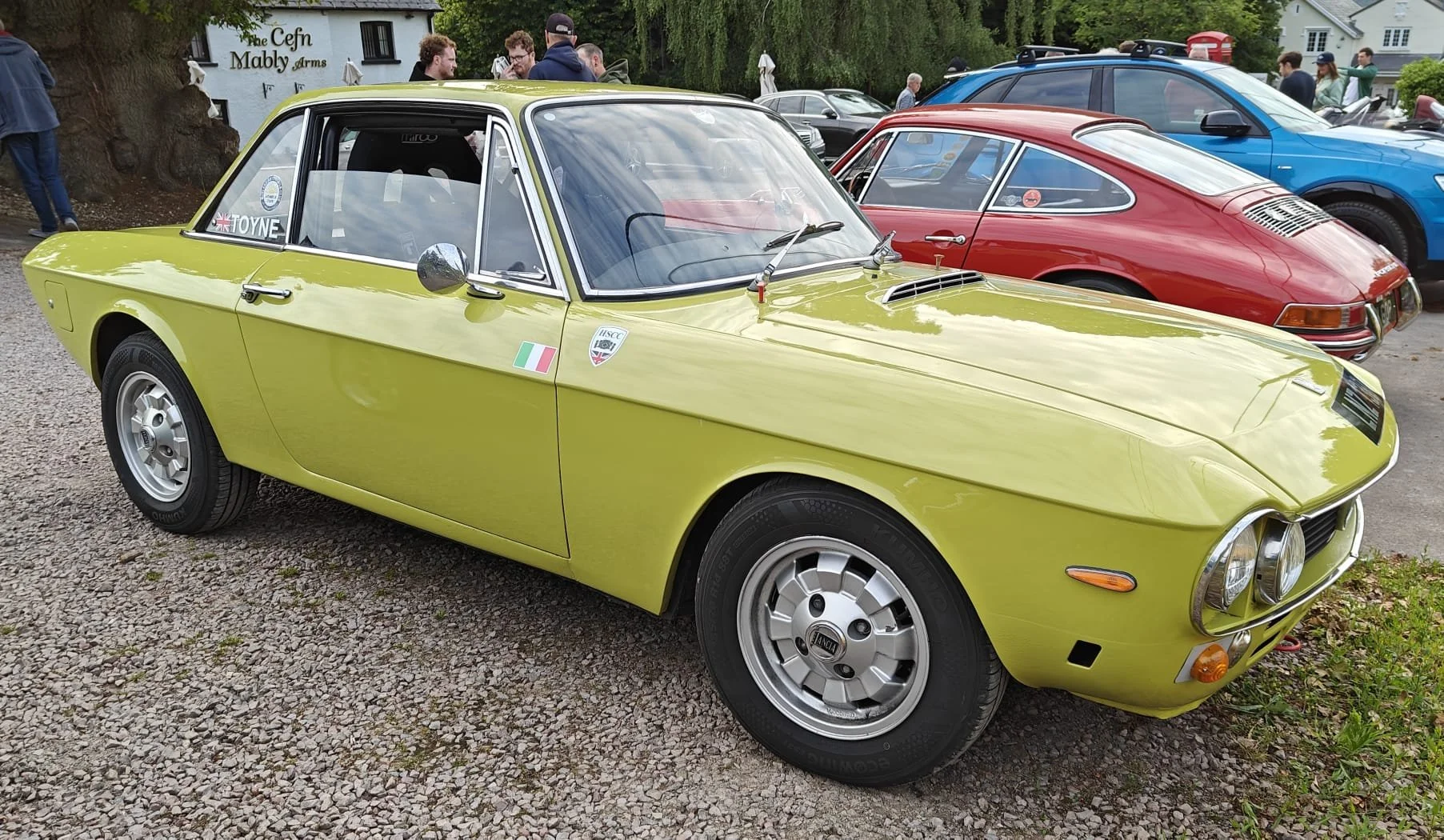 A vintage yellow-green sports car parked at a car show, with other classic cars and people in the background.