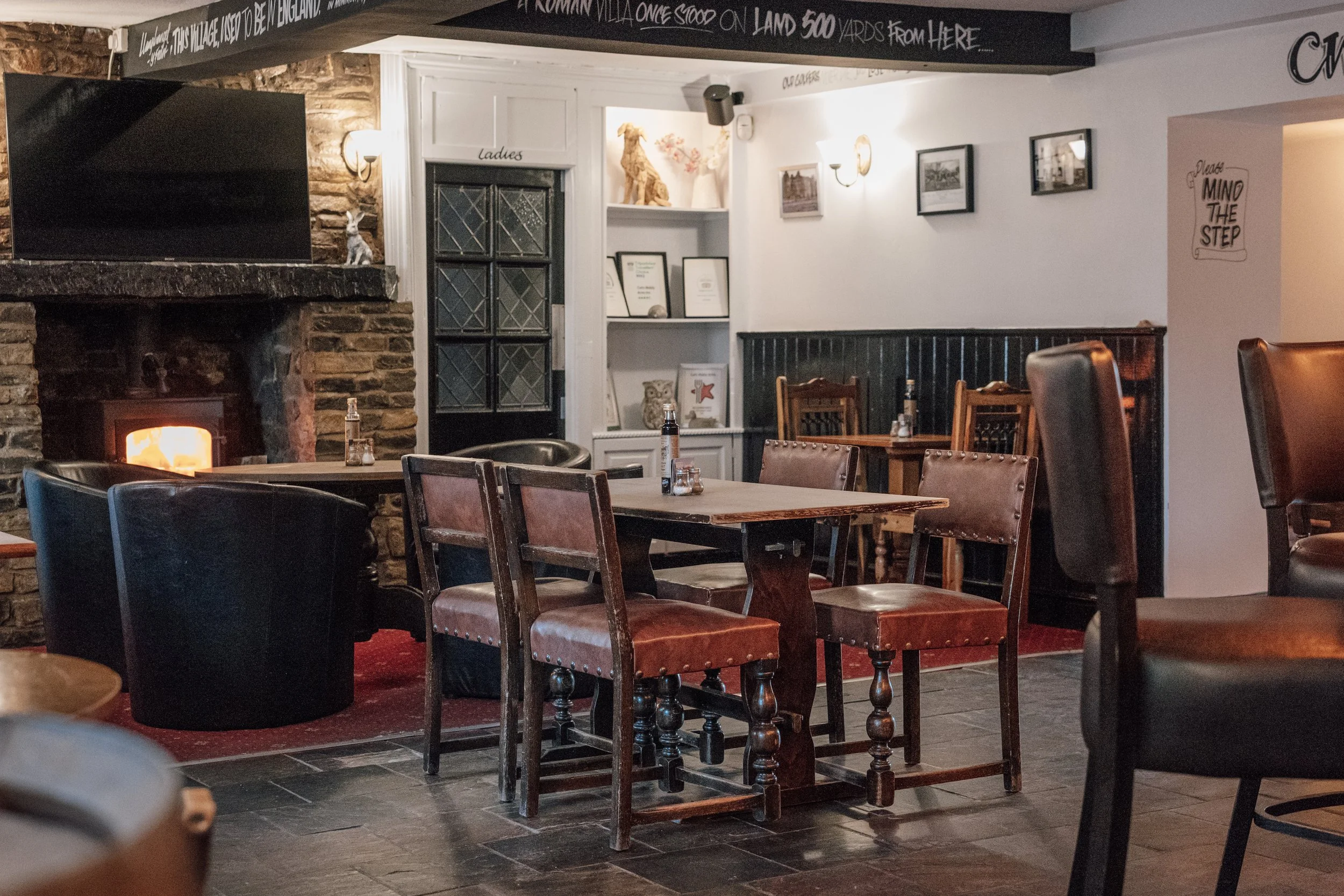 Dining area with wooden tables and chairs, black leather armchairs, a stone fireplace with a visible fire, framed pictures on the wall, bookshelves with framed certificates, and a decorative dog statue on the shelf.