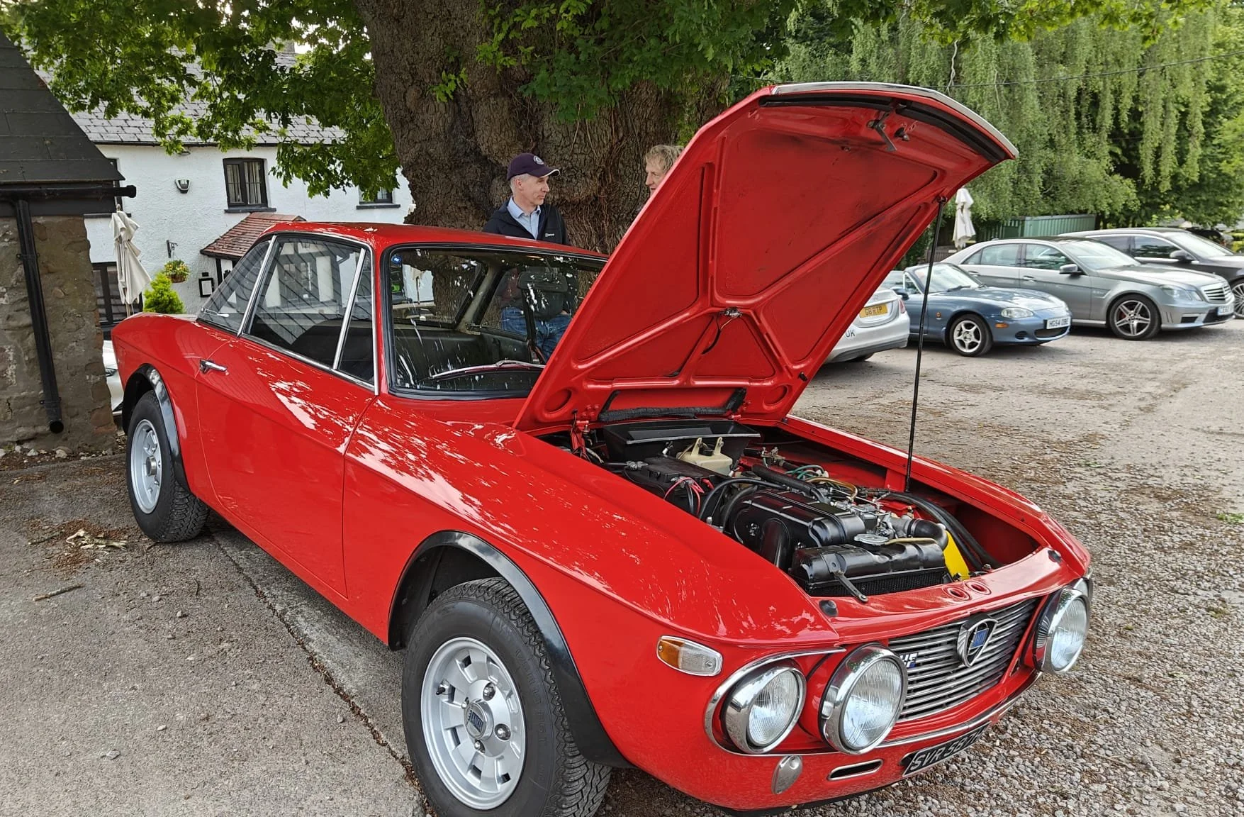 A red vintage car with its hood open, parked on a gravel surface, with two people talking nearby and other cars in the background under trees.