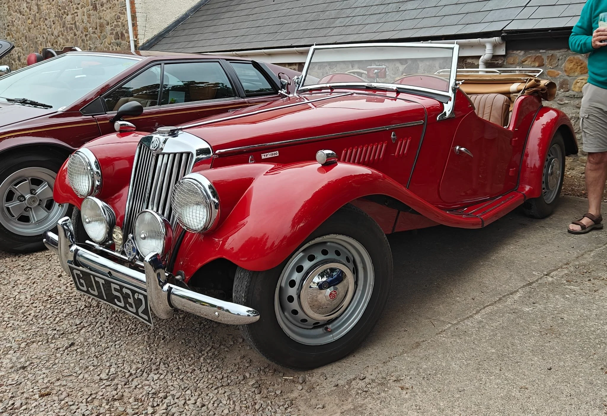 Red vintage MG TF 1500 convertible car parked on a gravel surface, with a gray modern car in the background and a person standing nearby.