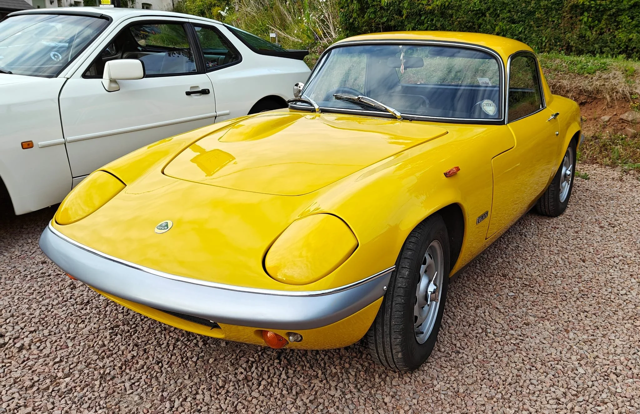 Yellow vintage Lotus Elise sports car parked on a gravel surface next to a white modern sports car with a background of green bushes and trees.