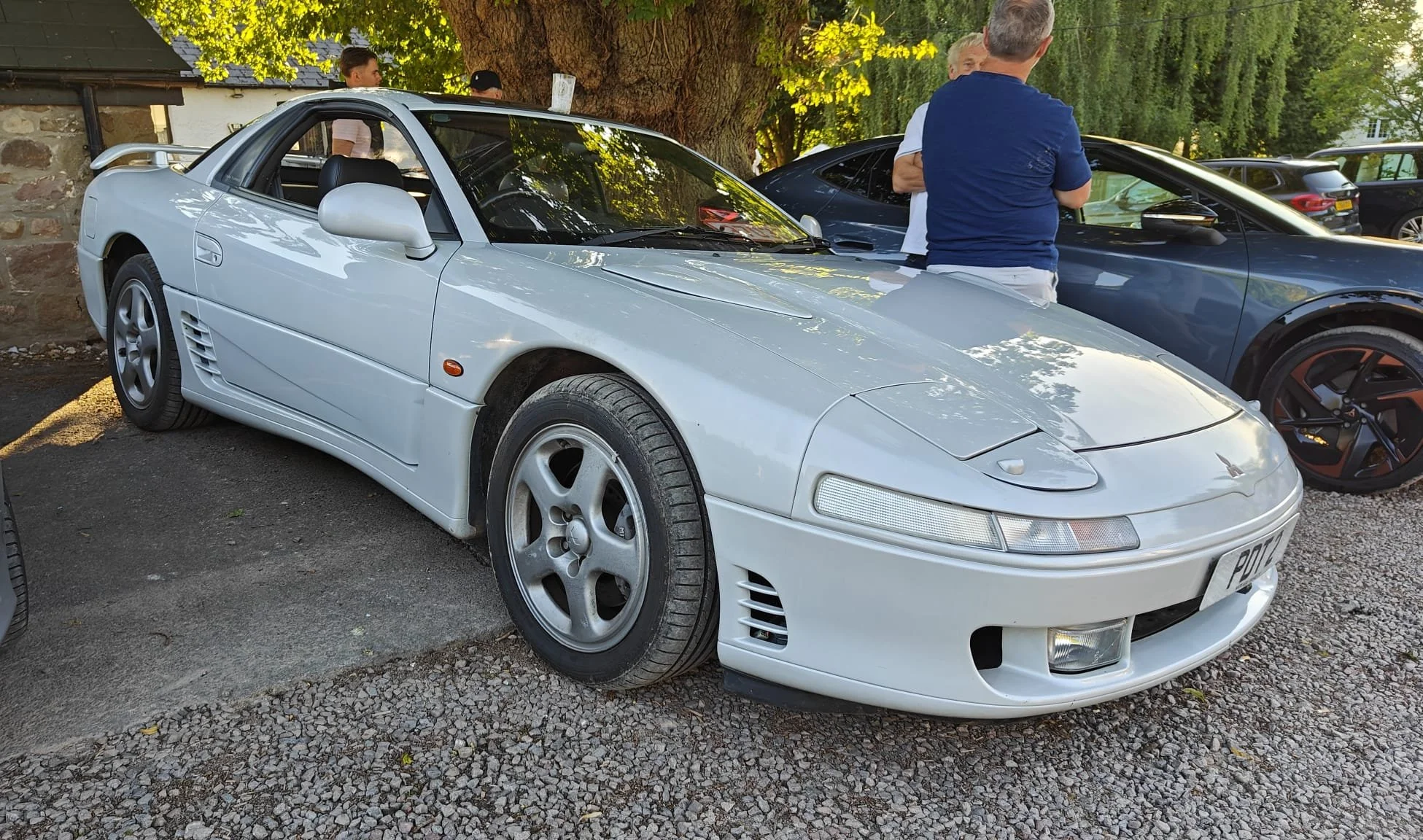 A white vintage sports car parked outdoors with several people in the background, next to a large tree.