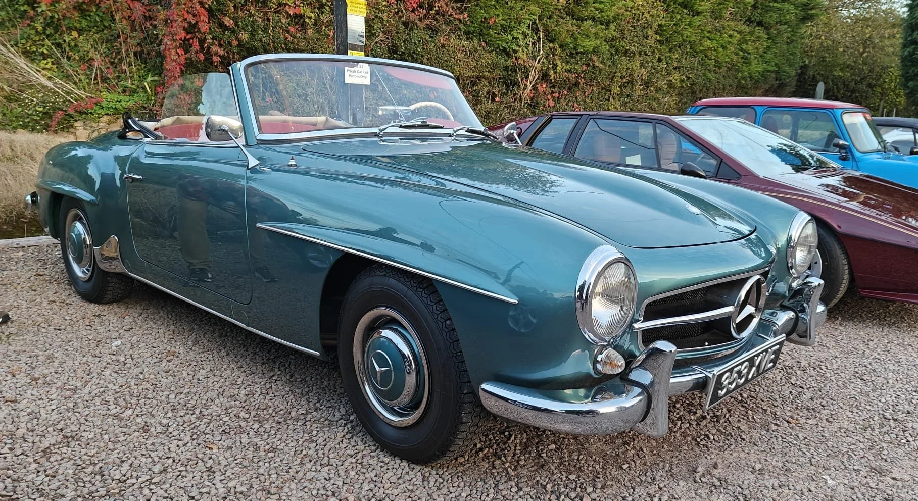 A vintage green Mercedes-Benz convertible sports car with chrome accents, parked among other classic cars, on a gravel surface with trees in the background.