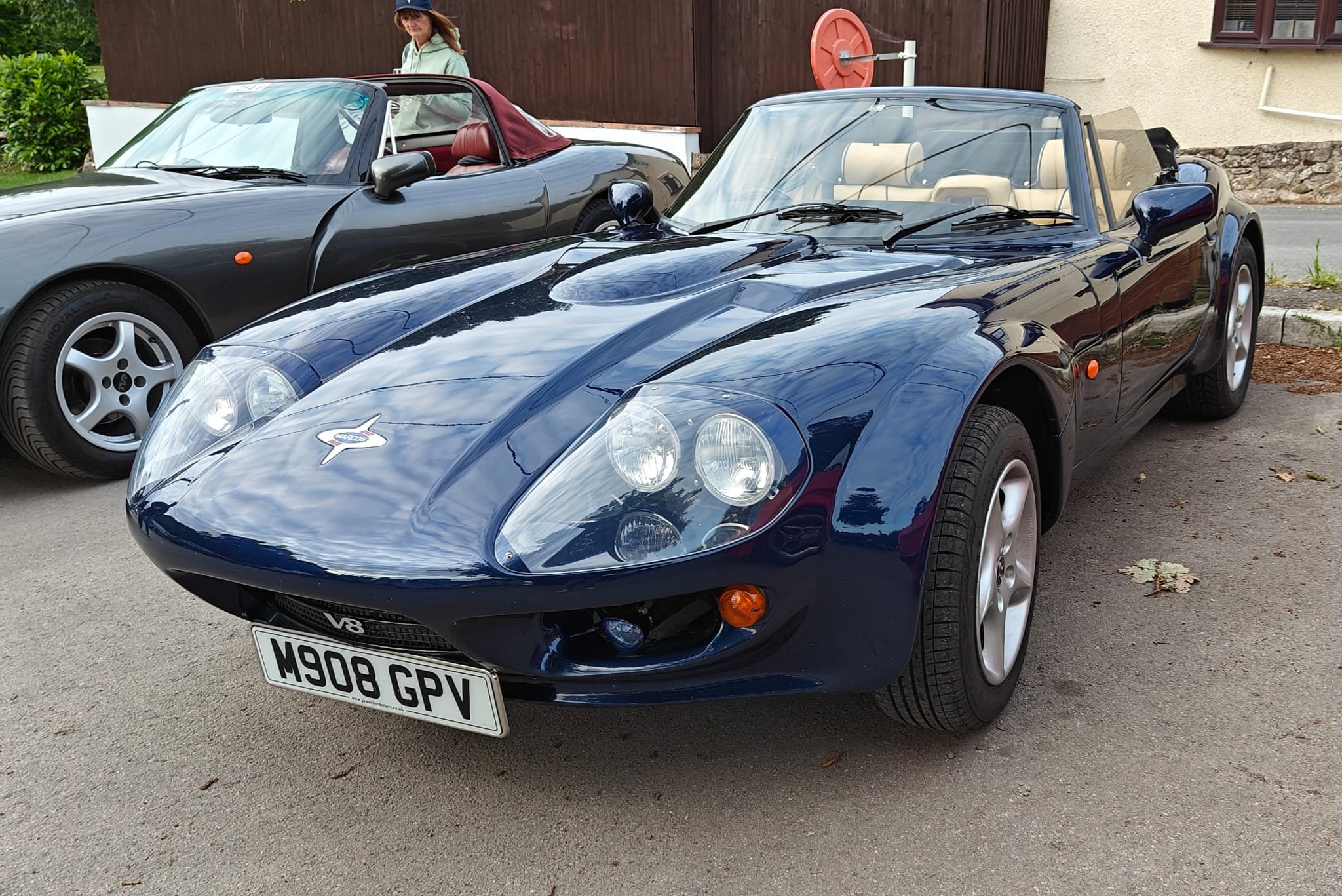 A dark blue vintage convertible sports car with beige interior, parked on a driveway next to a dark-colored convertible car. A woman standing behind the cars is looking at them, with a brown wooden building and a house in the background.