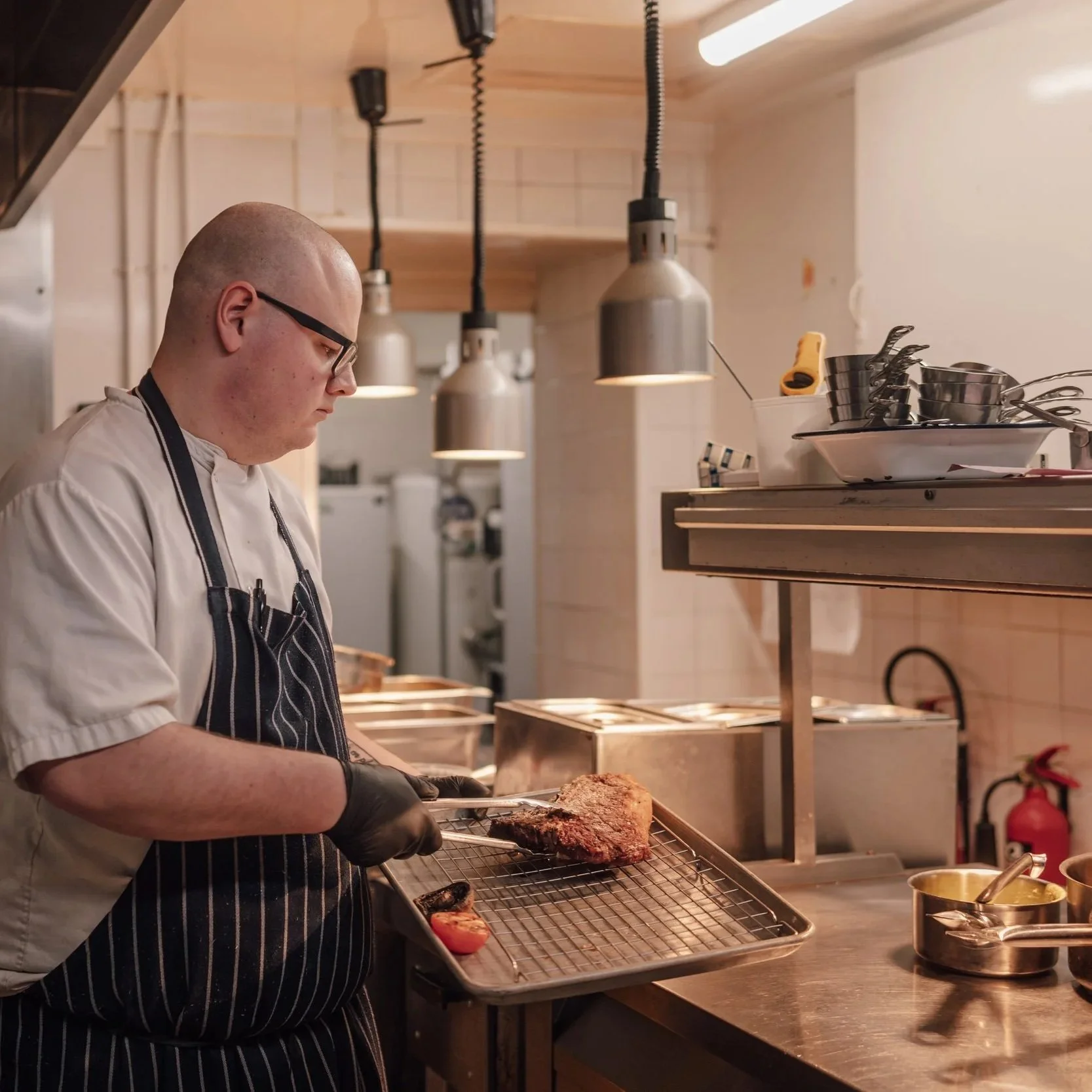 A chef wearing glasses, a striped apron, and black gloves is preparing a large piece of cooked meat on a wire rack in a professional kitchen.