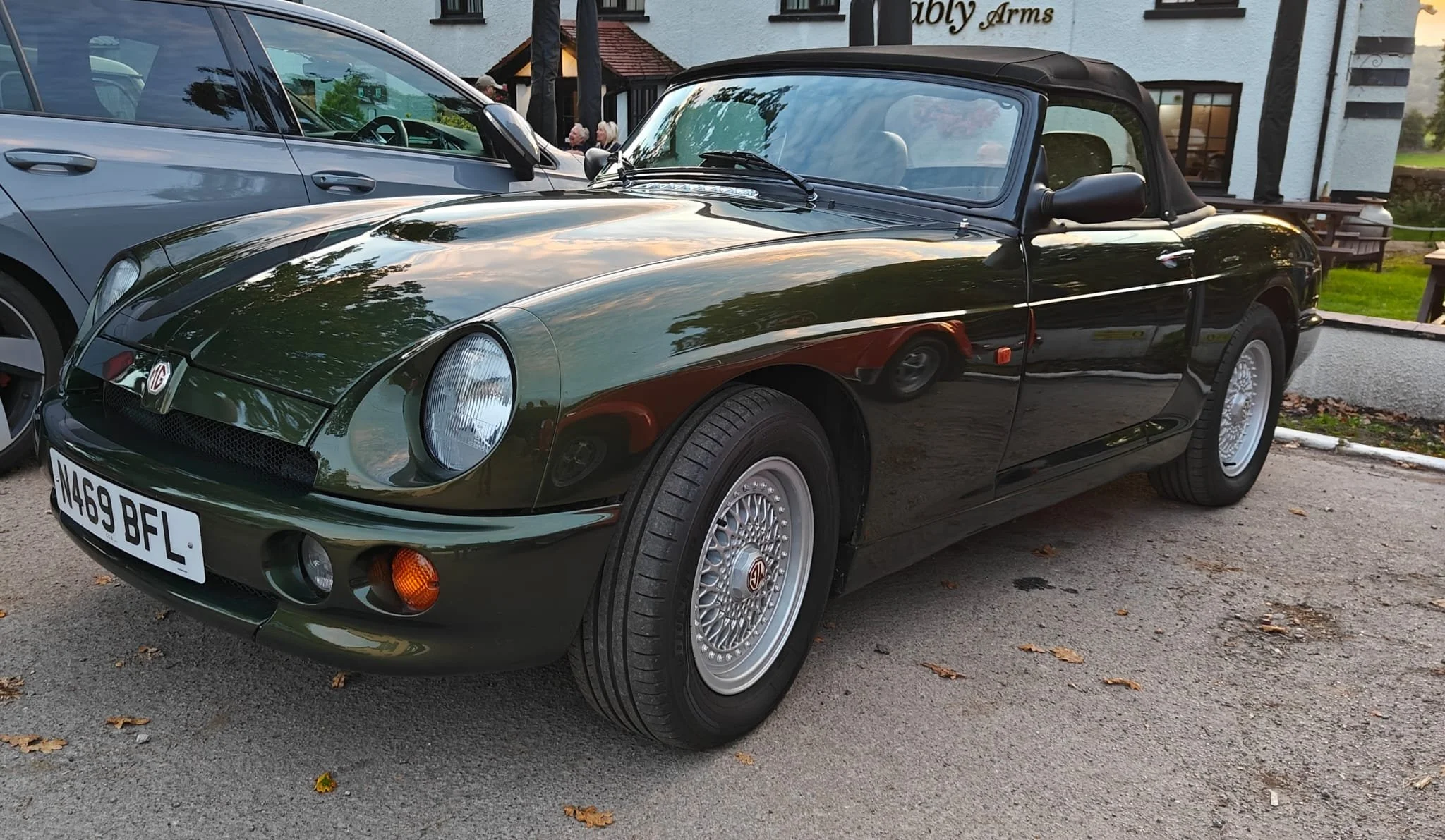 A classic dark green MG convertible sports car parked next to a modern silver sedan on a parking lot, with a building and trees reflected on the car's shiny surface.