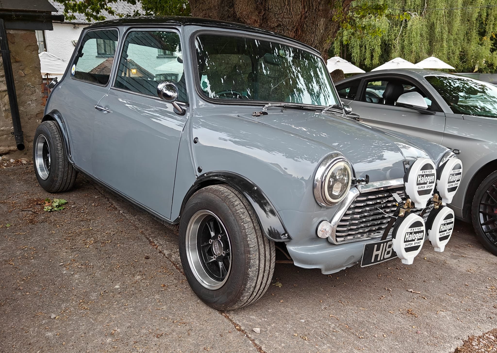 A vintage gray Mini Cooper with four fog lights mounted on the front grille, parked on a gravel surface, with another modern car next to it and trees in the background.