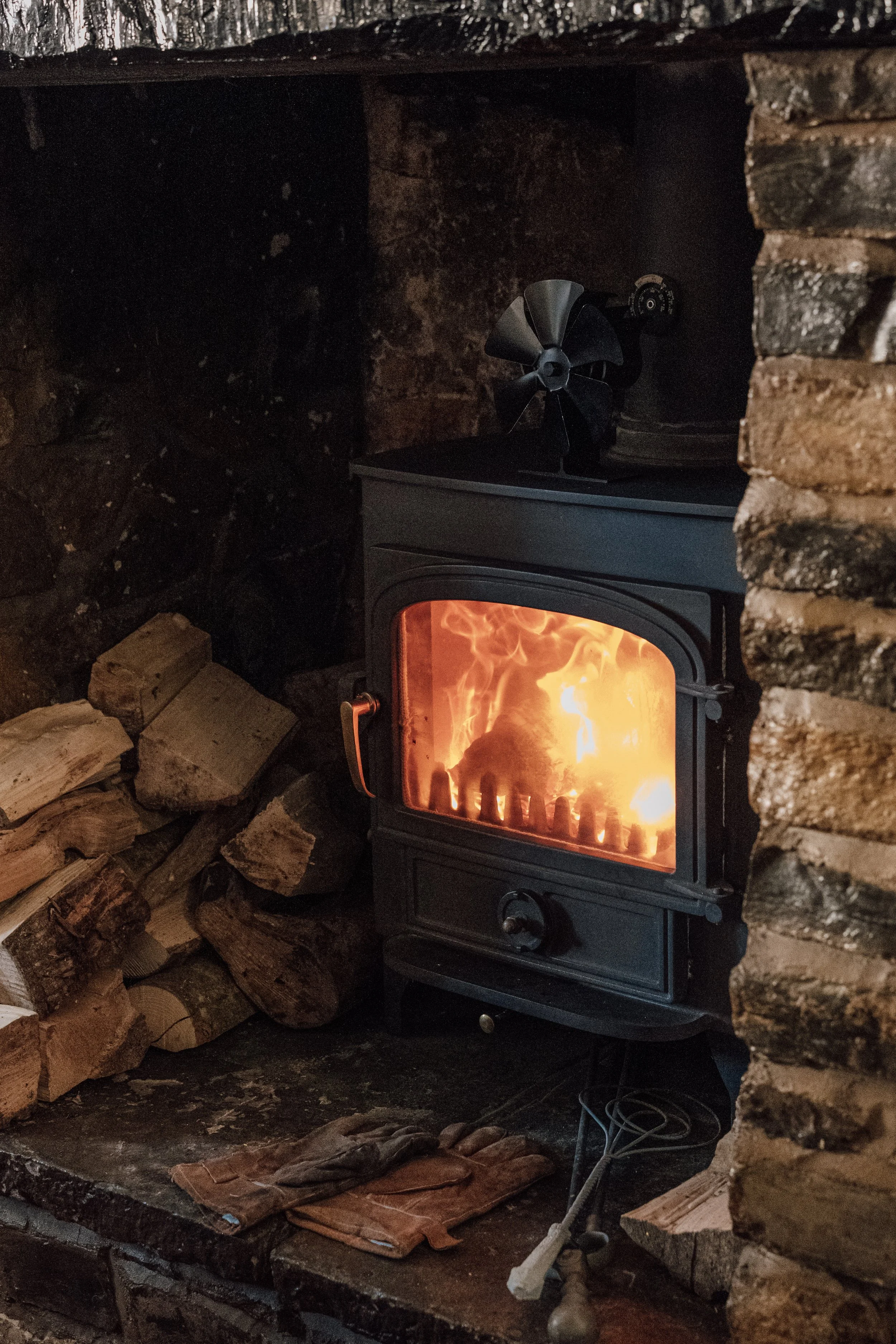 A wood-burning stove with a fire inside, surrounded by stacked firewood and a pair of gloves on the hearth, in a cozy rustic setting.