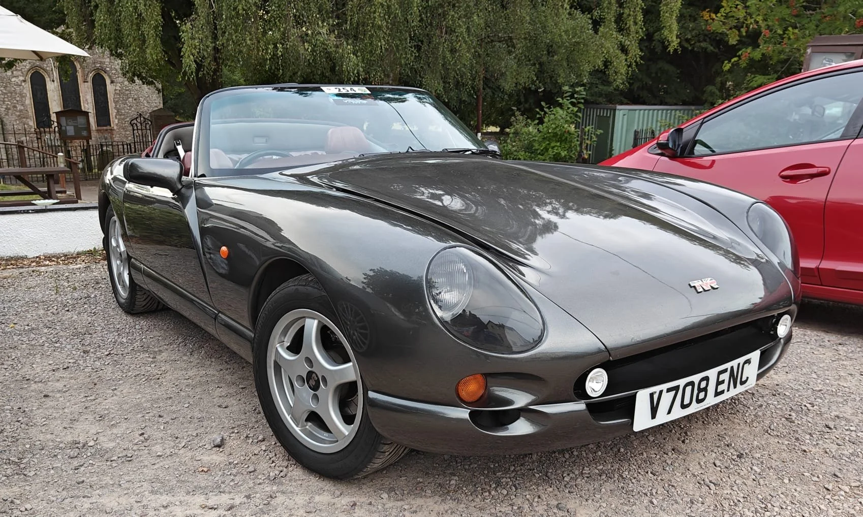 A classic black convertible sports car parked outdoors with a red car next to it. The car has rounded headlights, a sleek design, and a UK license plate.