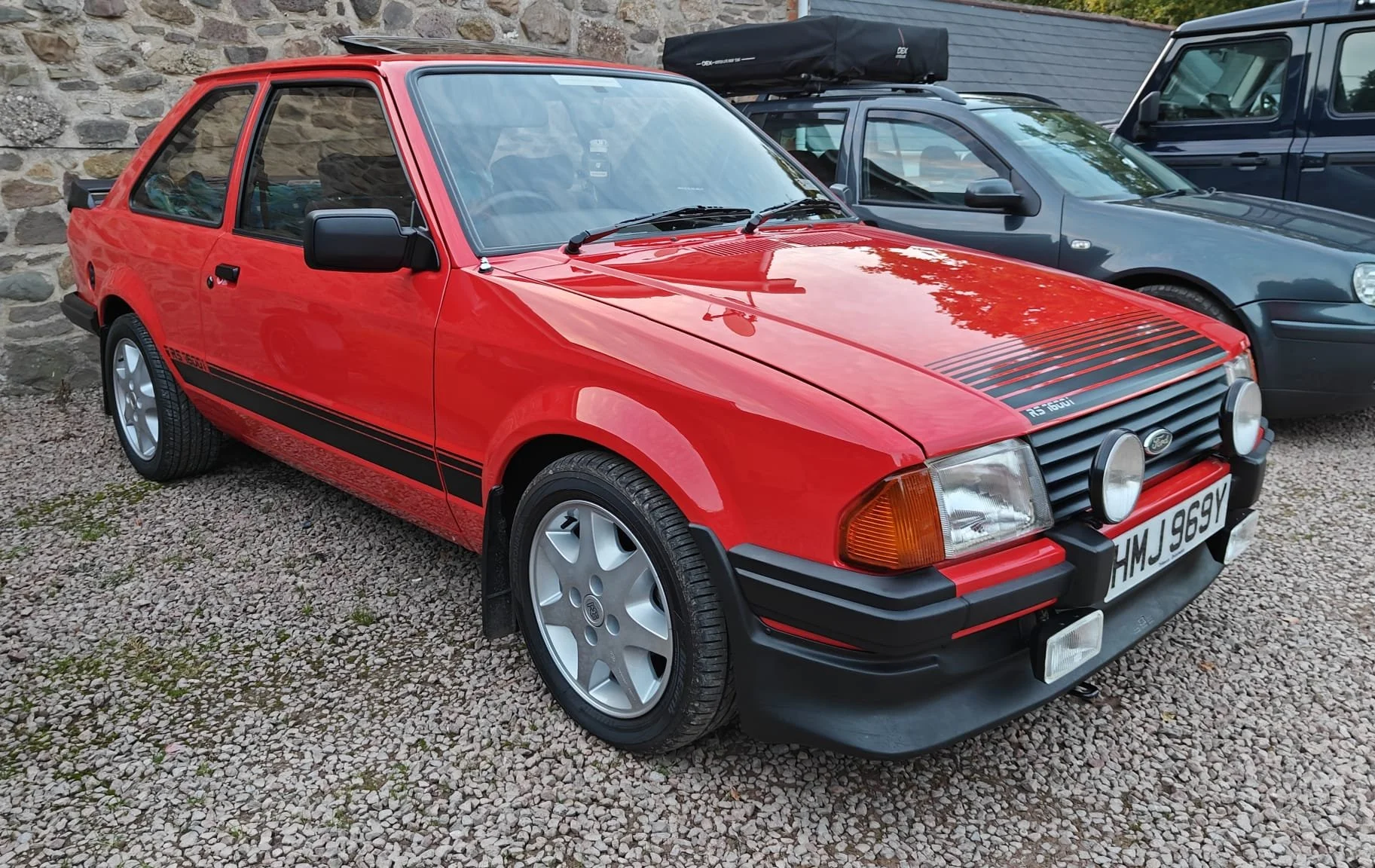A red vintage Ford Escort RS 1800 parked on gravel, with black racing stripes and fog lights, next to other cars.