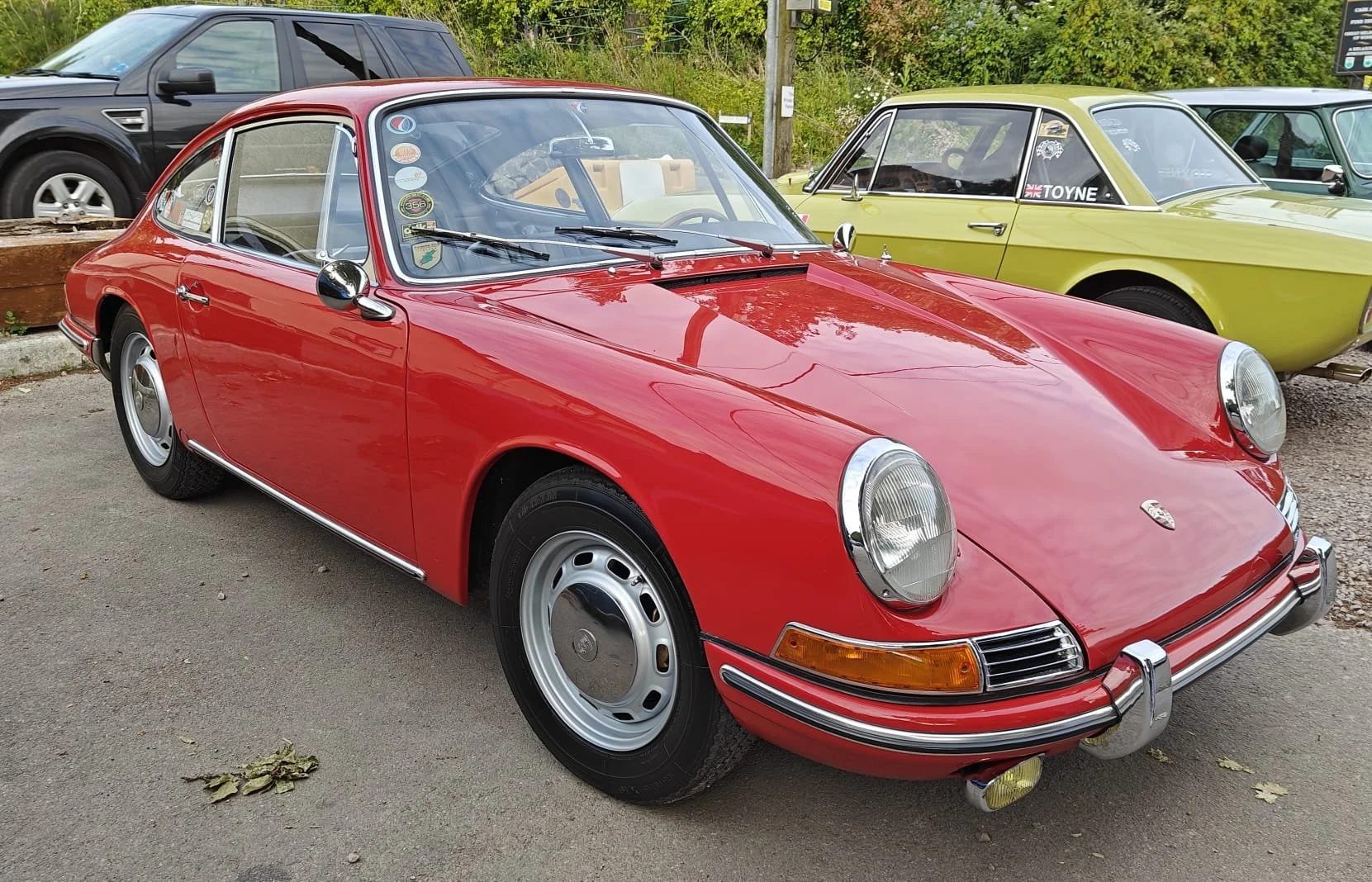 A vintage red Porsche 911 parked outdoors among other classic cars, with round headlights, chrome accents, and a distinctive sloped rear.