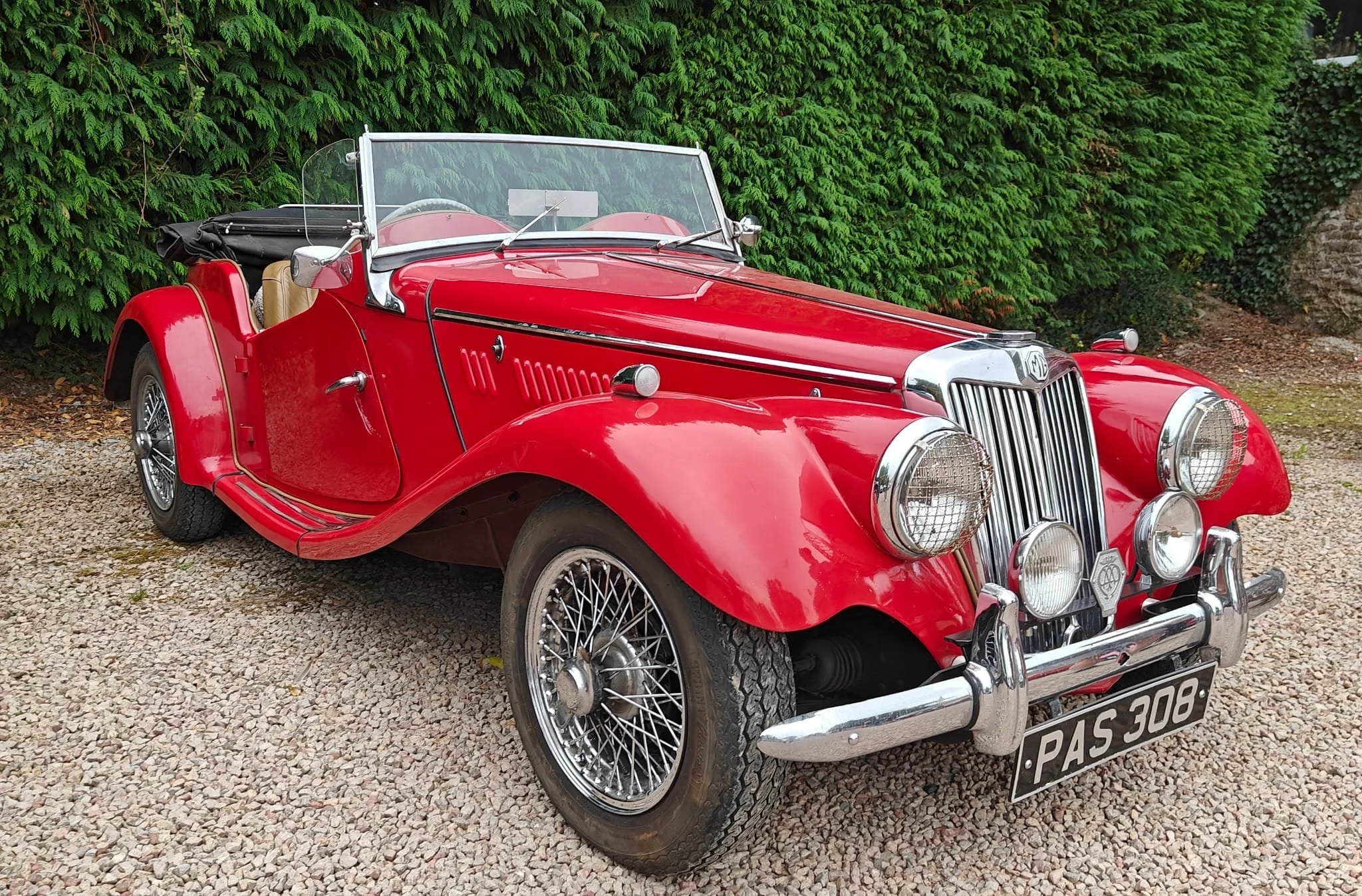 A vintage red convertible car parked on a gravel surface with green bushes in the background.