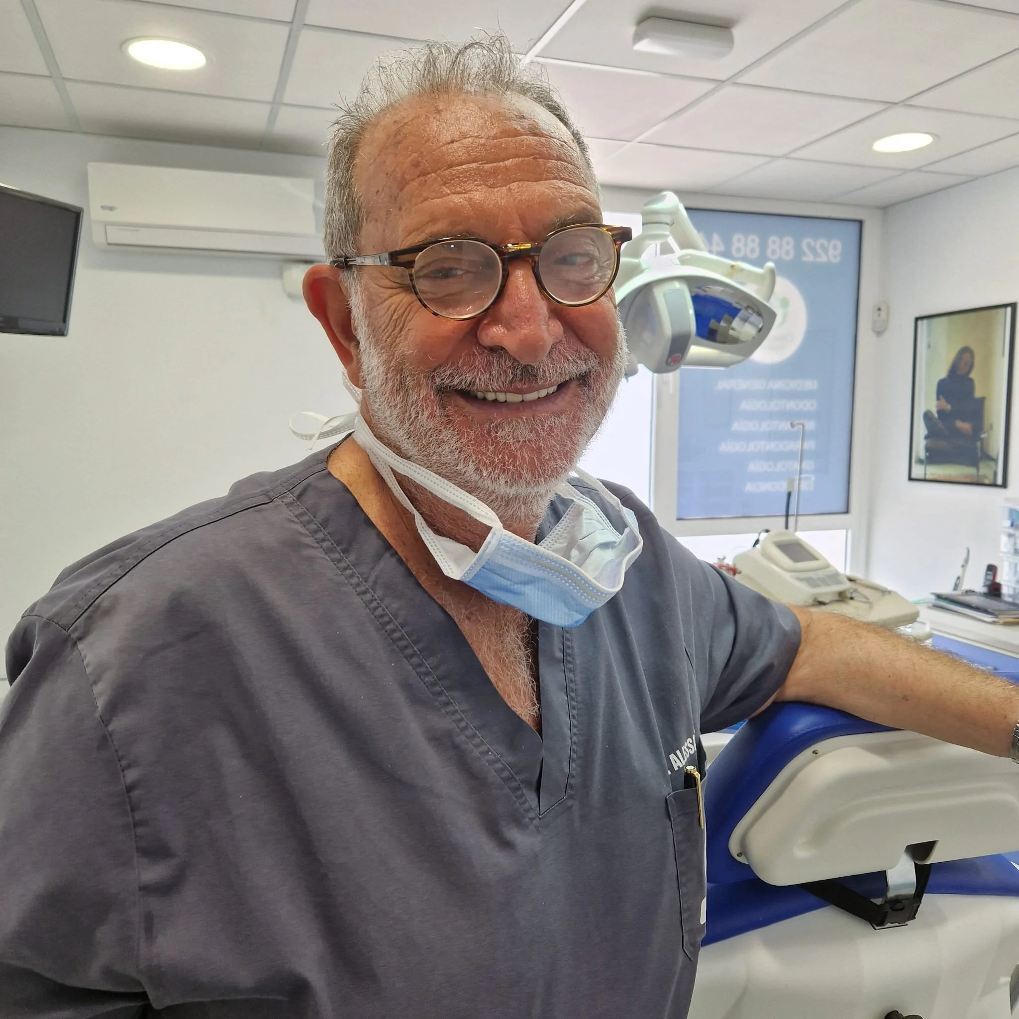 Un dentista sorridente con occhiali, camice grigia e mascherina abbassata, in uno studio dentistico moderno e ben illuminato.