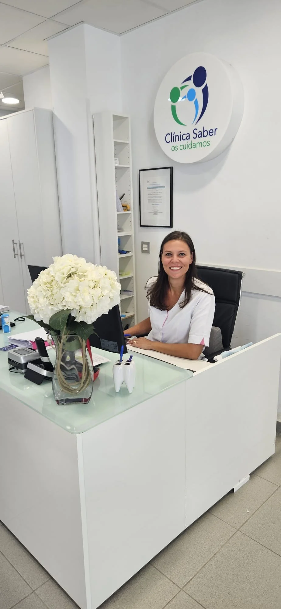 Una donna sorridente seduta dietro una scrivania in un ufficio, con un grande vaso di fiori bianchi di fronte a lei e uno schermo di computer. Sul muro dietro c'è un logo con il testo "Clínica Saber os cuidamos".