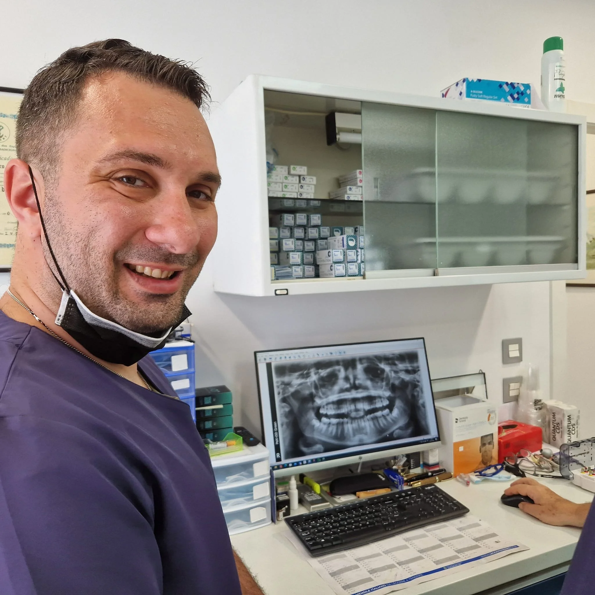 Un dentista sorridente con mascherina abbassata davanti a uno schermo con una radiografia dentale in uno studio dentistico.