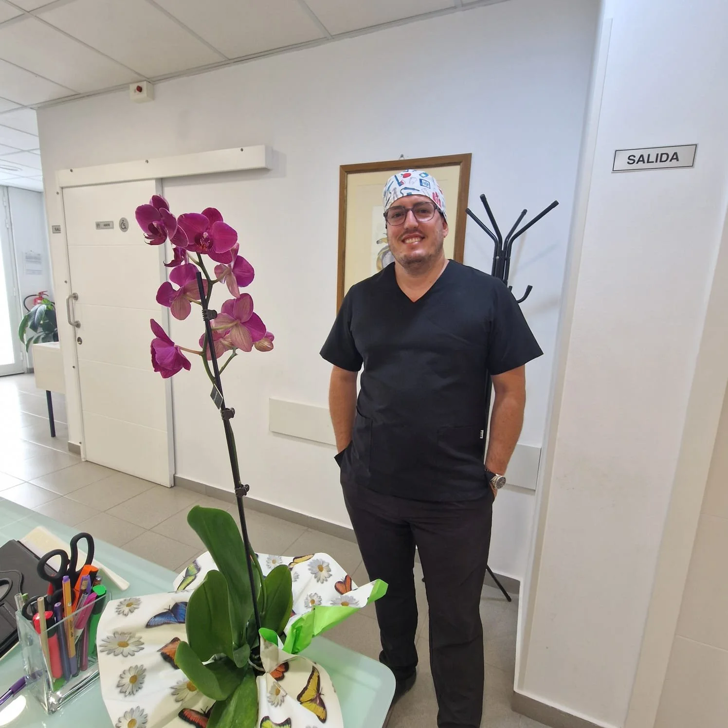 Un uomo sorridente vestito con divisa da medico o infermiere, con indosso una cuffia con disegni colorati, che si trova in un ambiente clinico o ospedaliero vicino a una pianta di orchidee rosa.