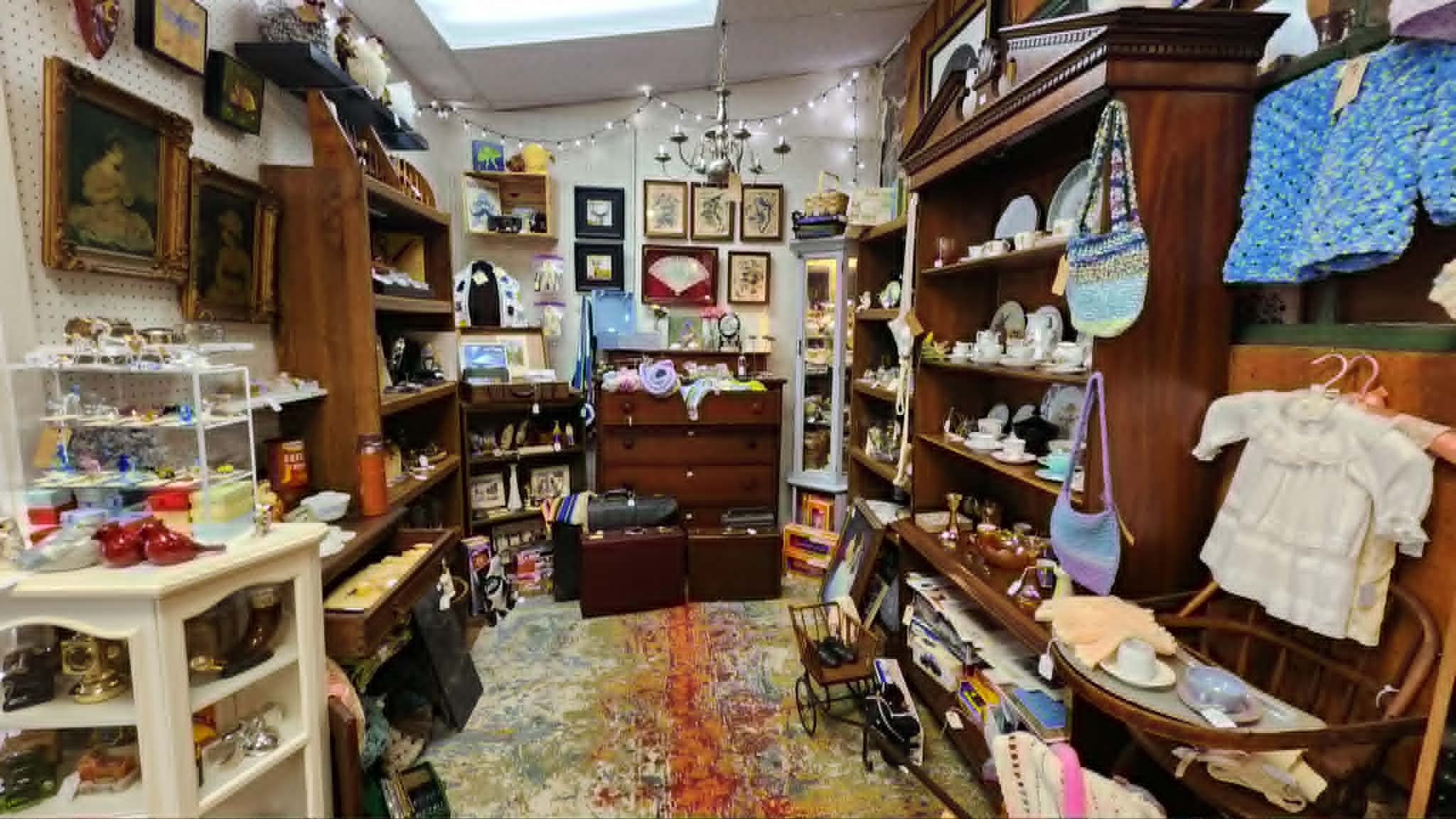 Inside a thrift store with shelves and tables filled with various knick-knacks, framed artwork, ceramics, and collectibles. There are string lights hanging from the ceiling, and a vintage rug on the floor.