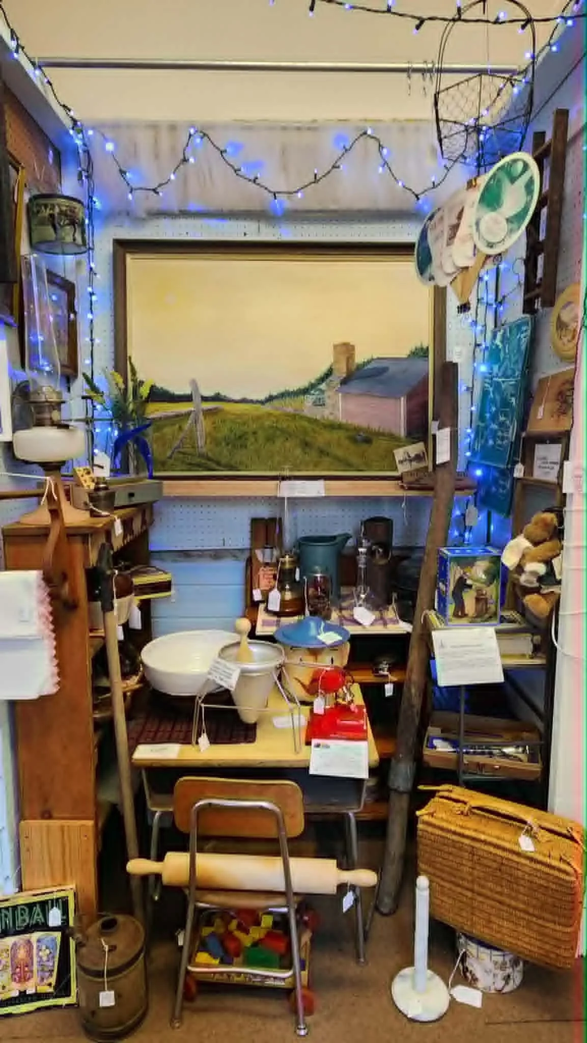 A thrift store display with various household and decorative items, including a large painting of a countryside scene, string lights, shelves with knickknacks, toys, and vintage items.
