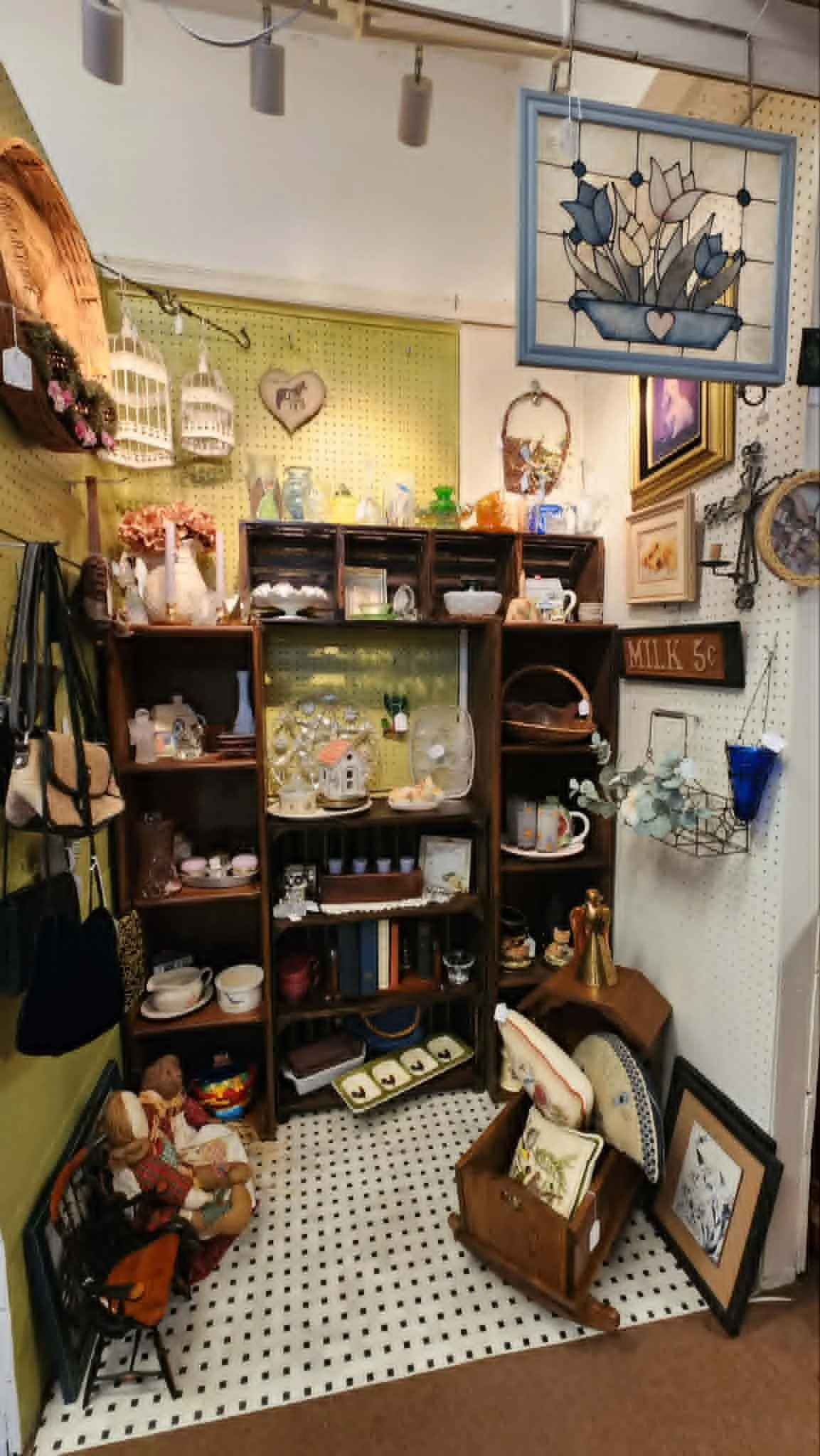 A small vintage thrift store display with various decorative items and collectibles. There are shelves filled with pottery, glassware, and small decorative objects, as well as framed artwork and a hanging stained glass panel. A doll in a plaid dress 