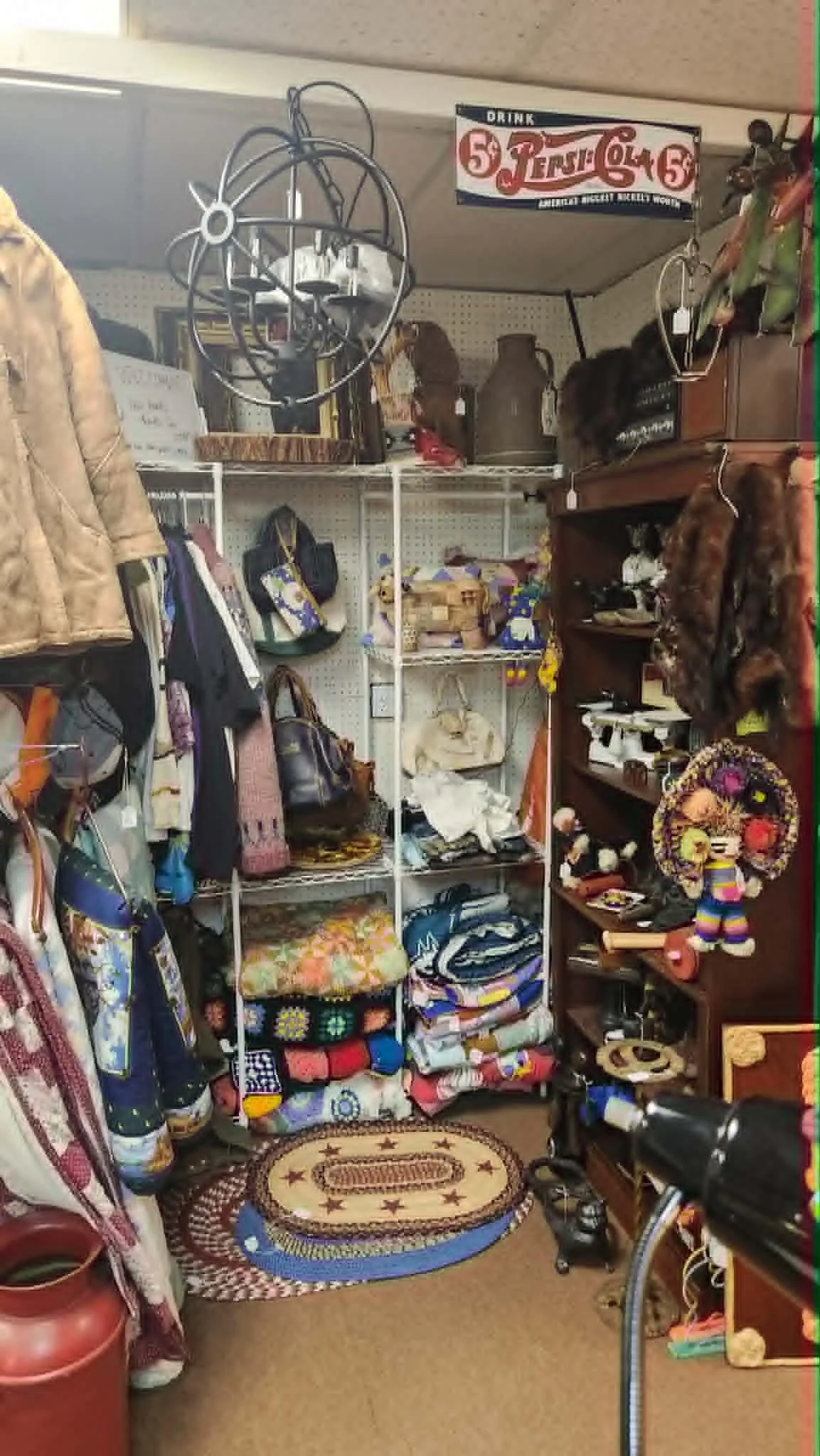 A thrift store aisle with shelves holding bags, toys, and decorative items, a coiled vacuum hose, and a bicycle. There are clothes hanging on the left side and a small rug on the floor with a tan carpet background.