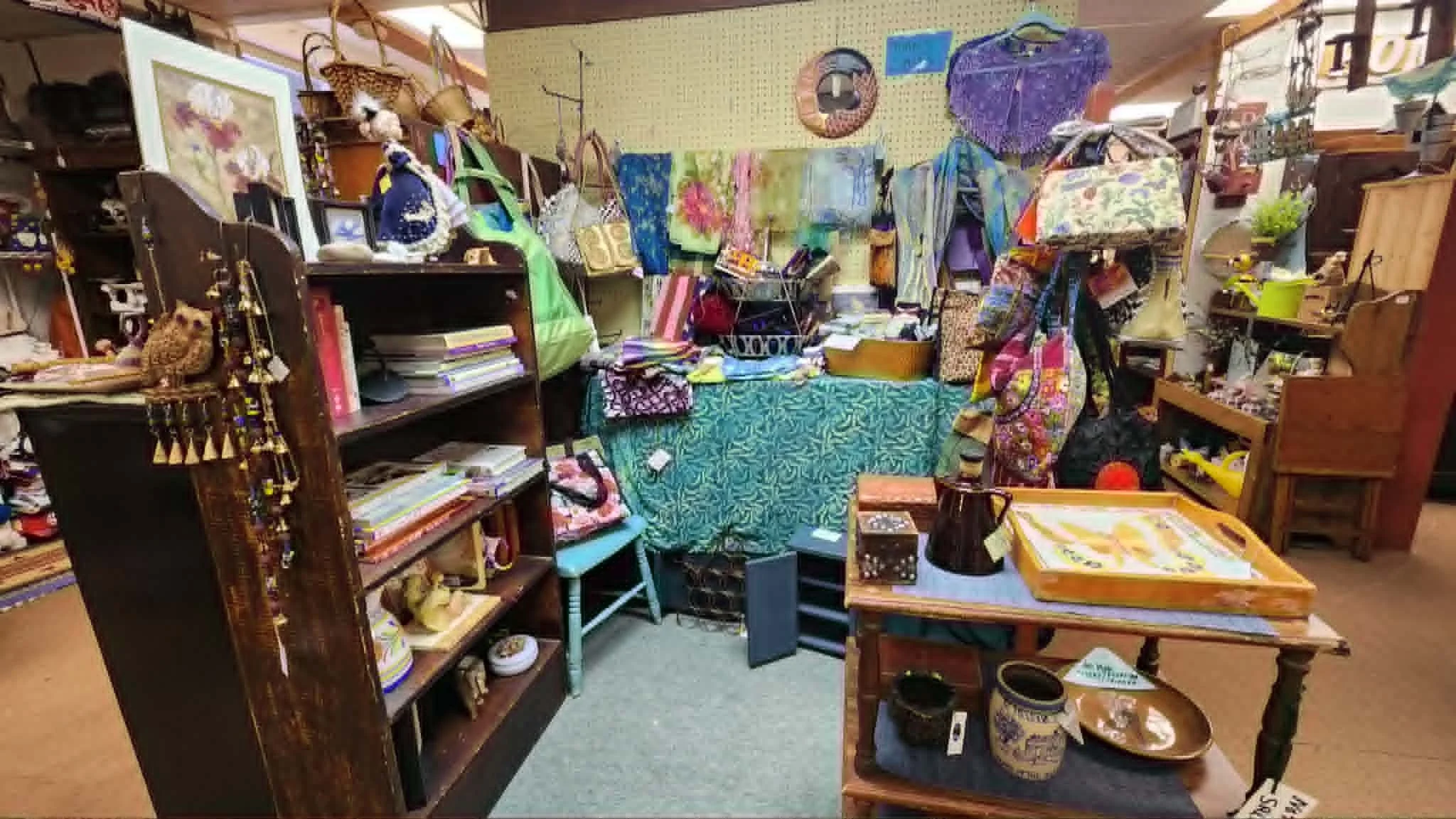 A cluttered thrift store or antique shop with various furniture, bags, and decorative items on shelves and tables, including picture frames, toys, and fabric pieces.