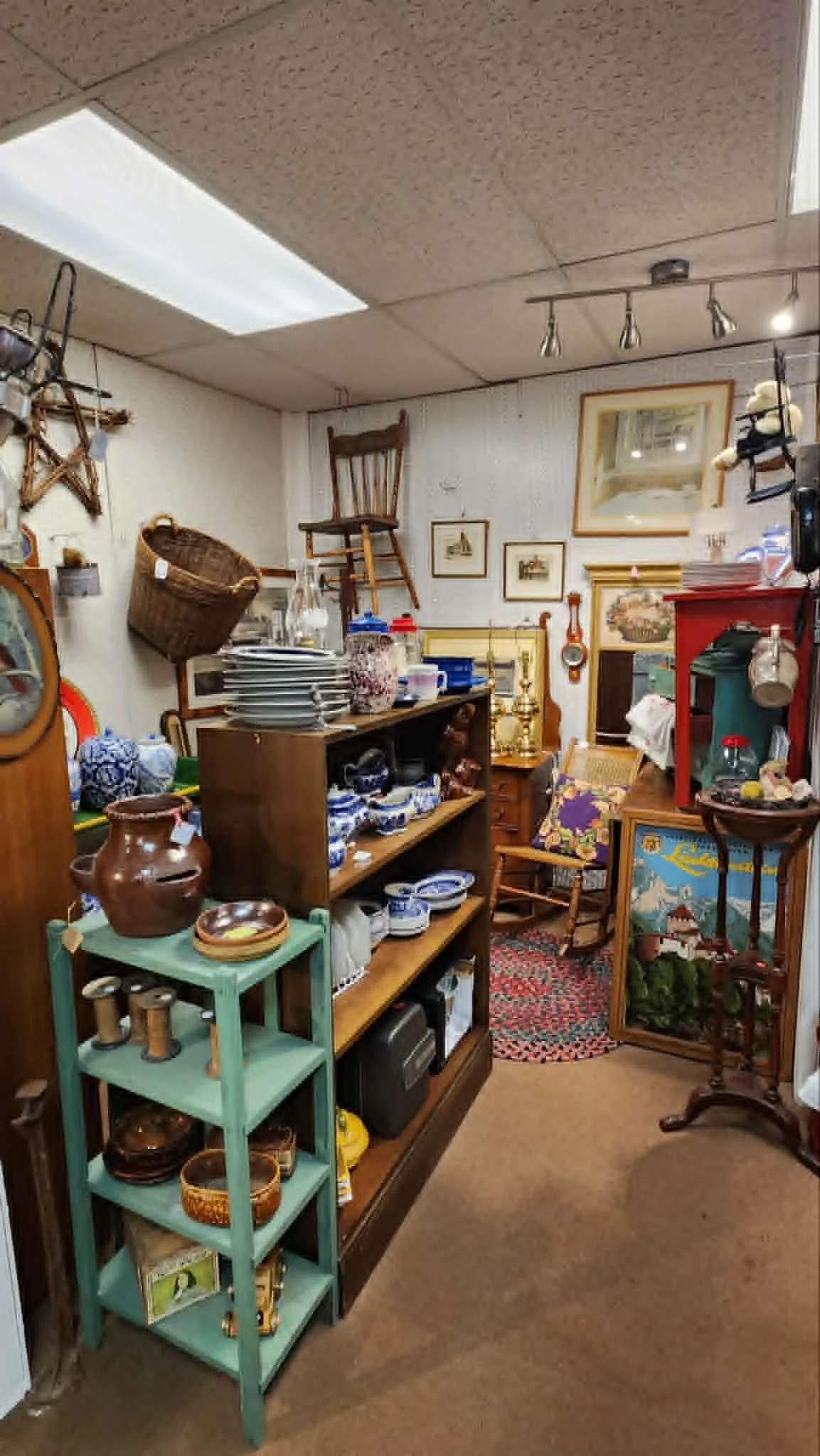 Interior of an antique shop with shelves of dishes, pottery, and decorative items. There is a wooden chair on the wall, frames, and a display case with collectibles. The ceiling has fluorescent and track lighting.