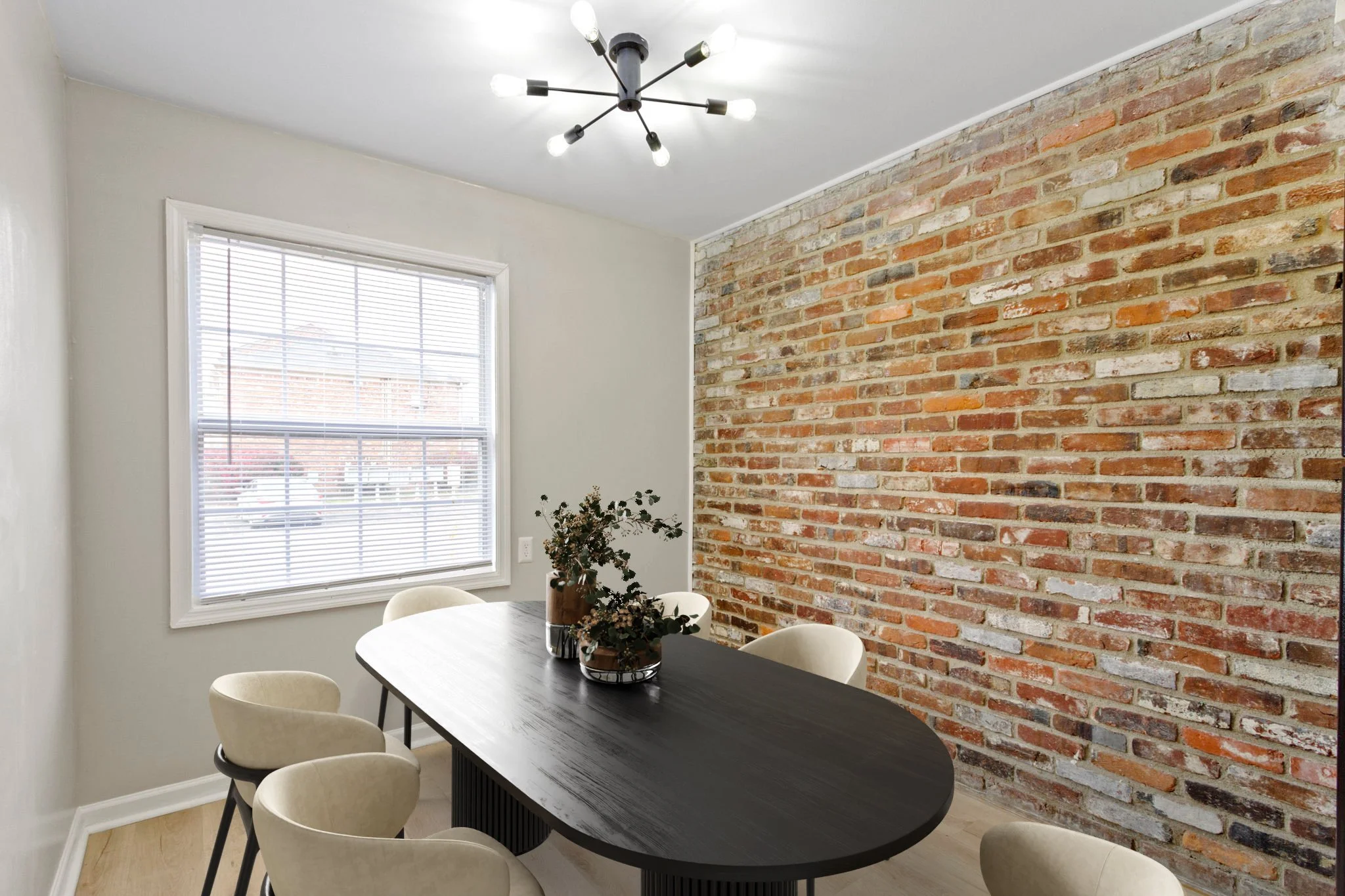 A dining room with a black oval table, six beige chairs, a brick accent wall, a large window with blinds, and a modern chandelier with multiple bulbs.