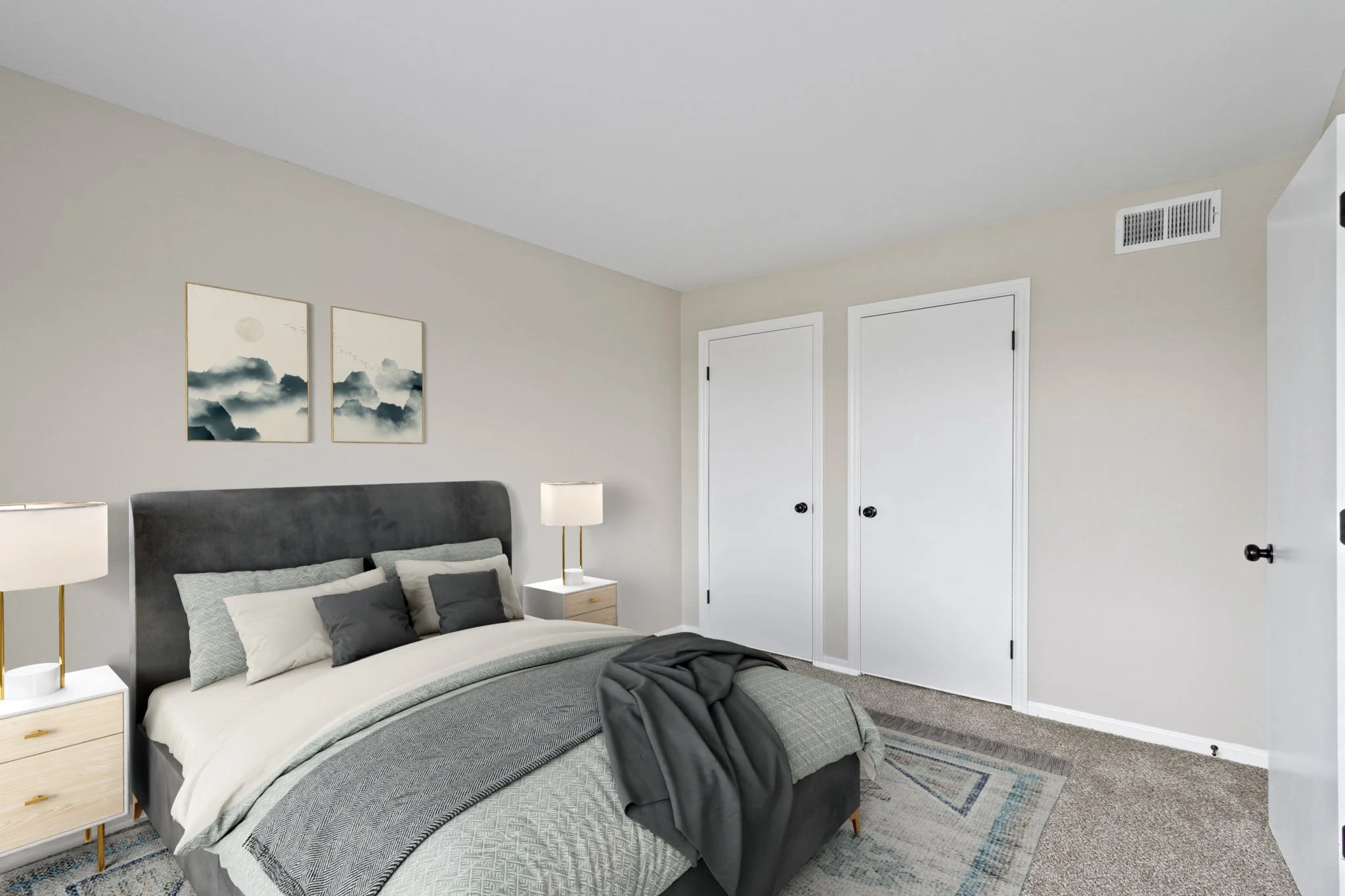 A neatly made bed with a dark gray headboard, gray and white bedding, and some gray pillows, in a bedroom with two nightstands and lamps. There are two framed art prints of mountains and a moon on the wall above the bed, beige walls, a carpeted floor