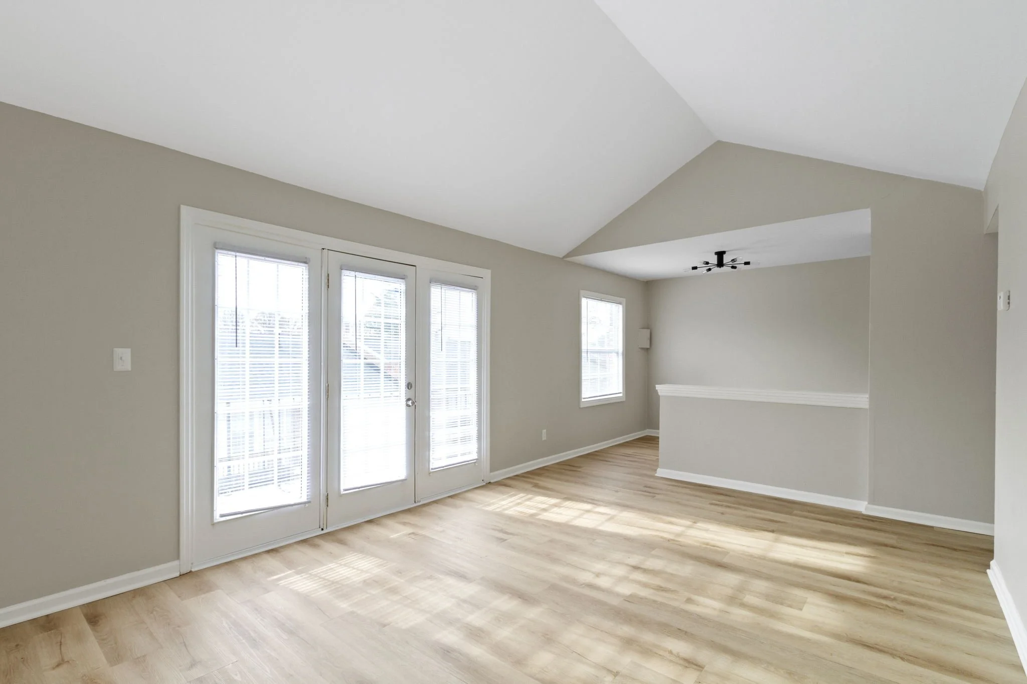 Empty living room with light-colored hardwood floors, white walls, and a ceiling with a modern black ceiling fan. There are three large windows with blinds and glass door leading outside.