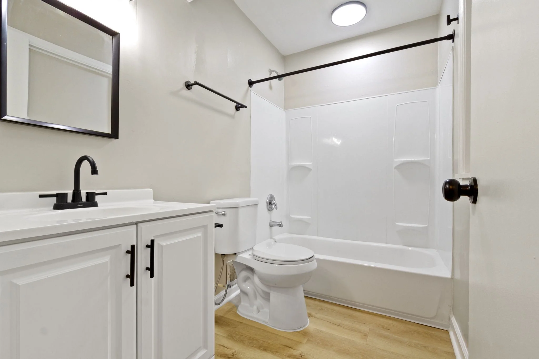 A small, clean bathroom with a white vanity, black hardware, and a large mirror. There is a toilet next to the vanity. The bathtub and shower combination has a black shower rod and a white surround with built-in shelves. The flooring is light wood, a