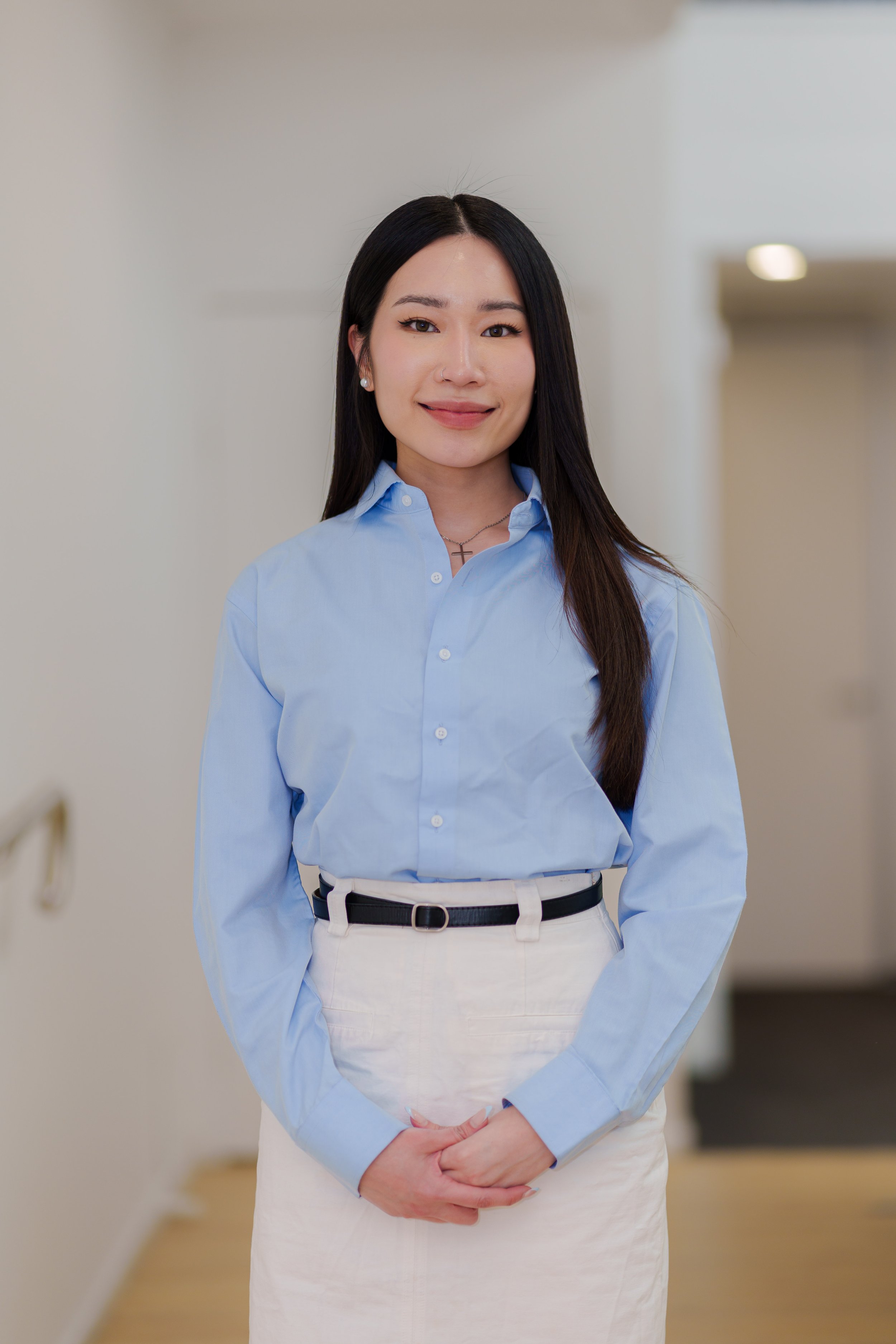 Portrait of a young Asian woman with long black hair, wearing a light blue button-down shirt and white skirt, standing indoors with her hands clasped in front of her.