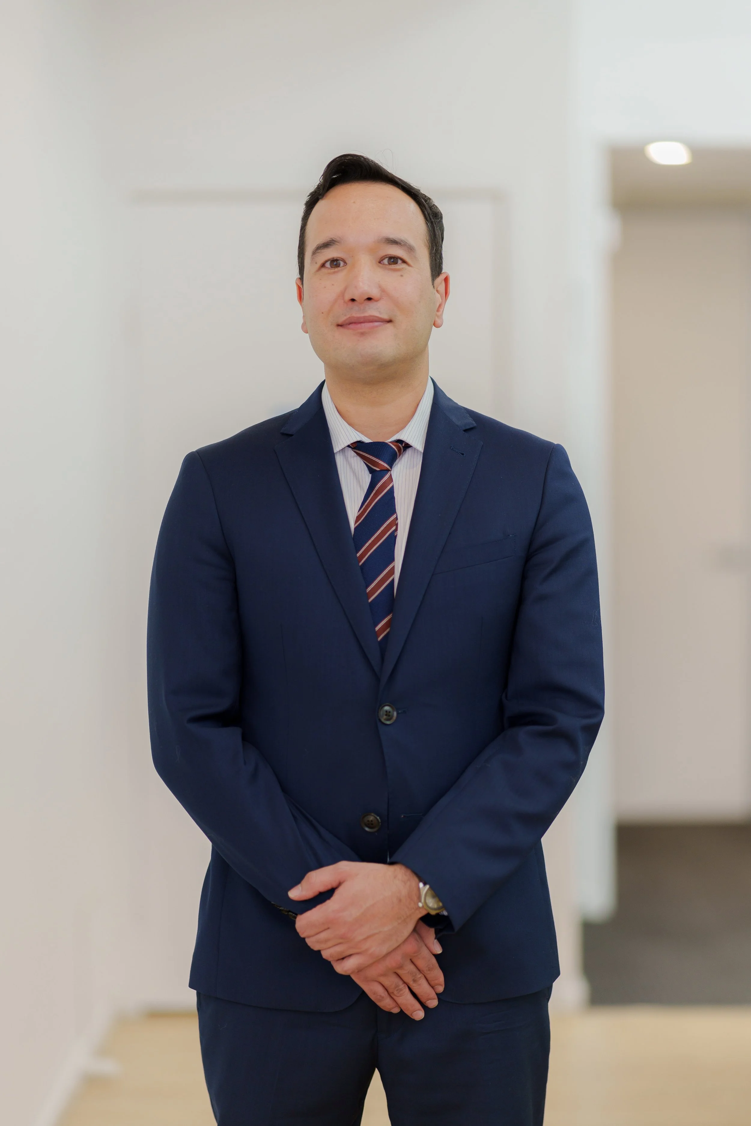 A man in a navy blue suit with a collared shirt and striped tie, standing indoors in front of a plain light-colored wall with soft lighting.