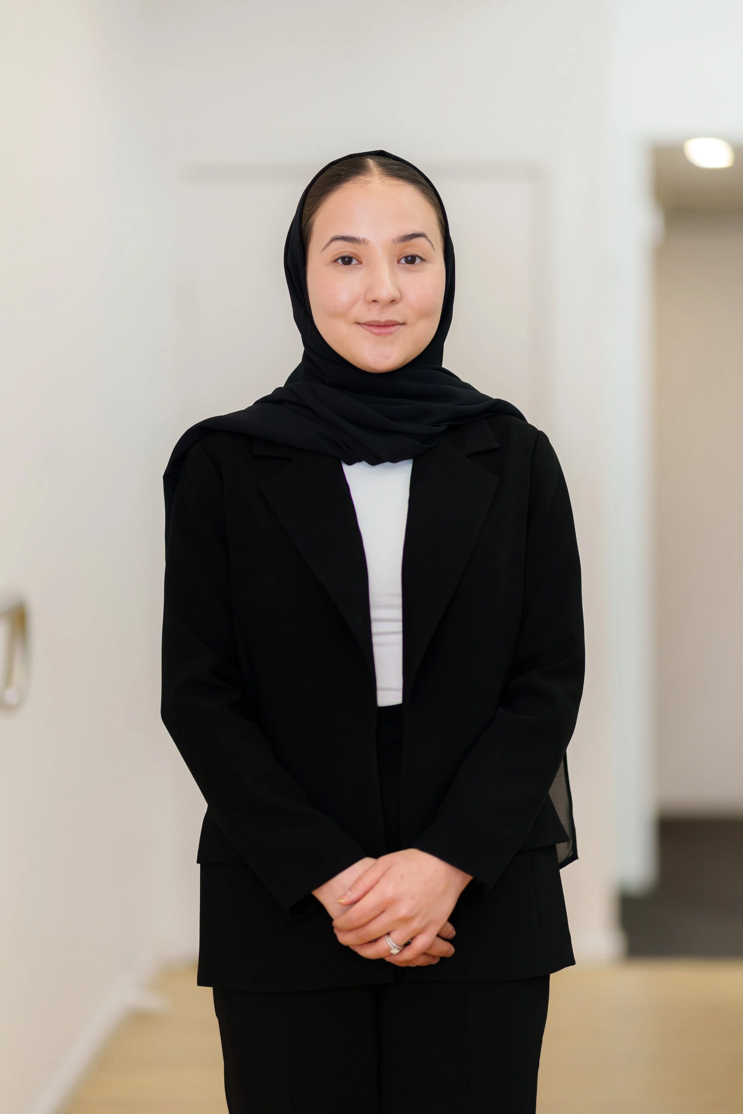 A woman in a black blazer and hijab standing in a neutral indoor setting, smiling gently.