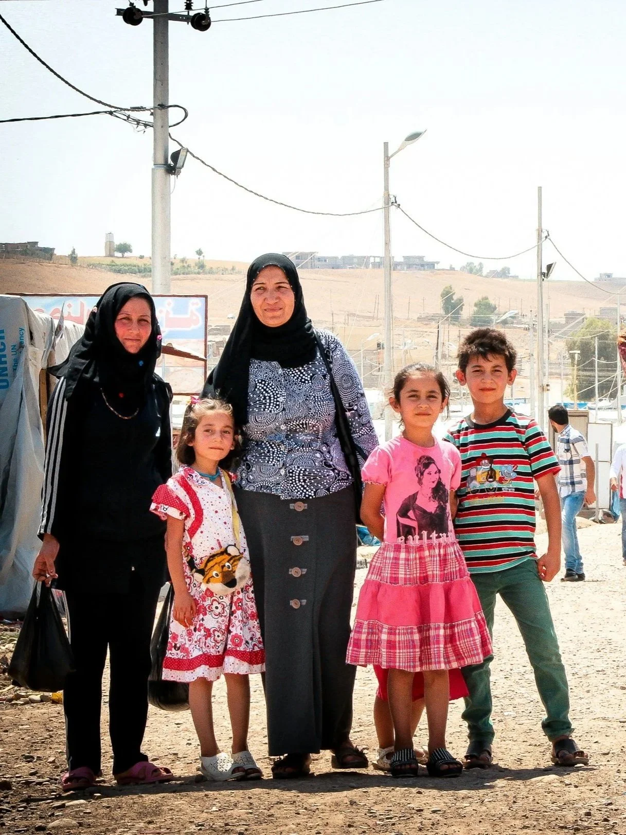 A group of six people, two women and four children, standing outdoors in a sunny, rural area with hills and power lines in the background.