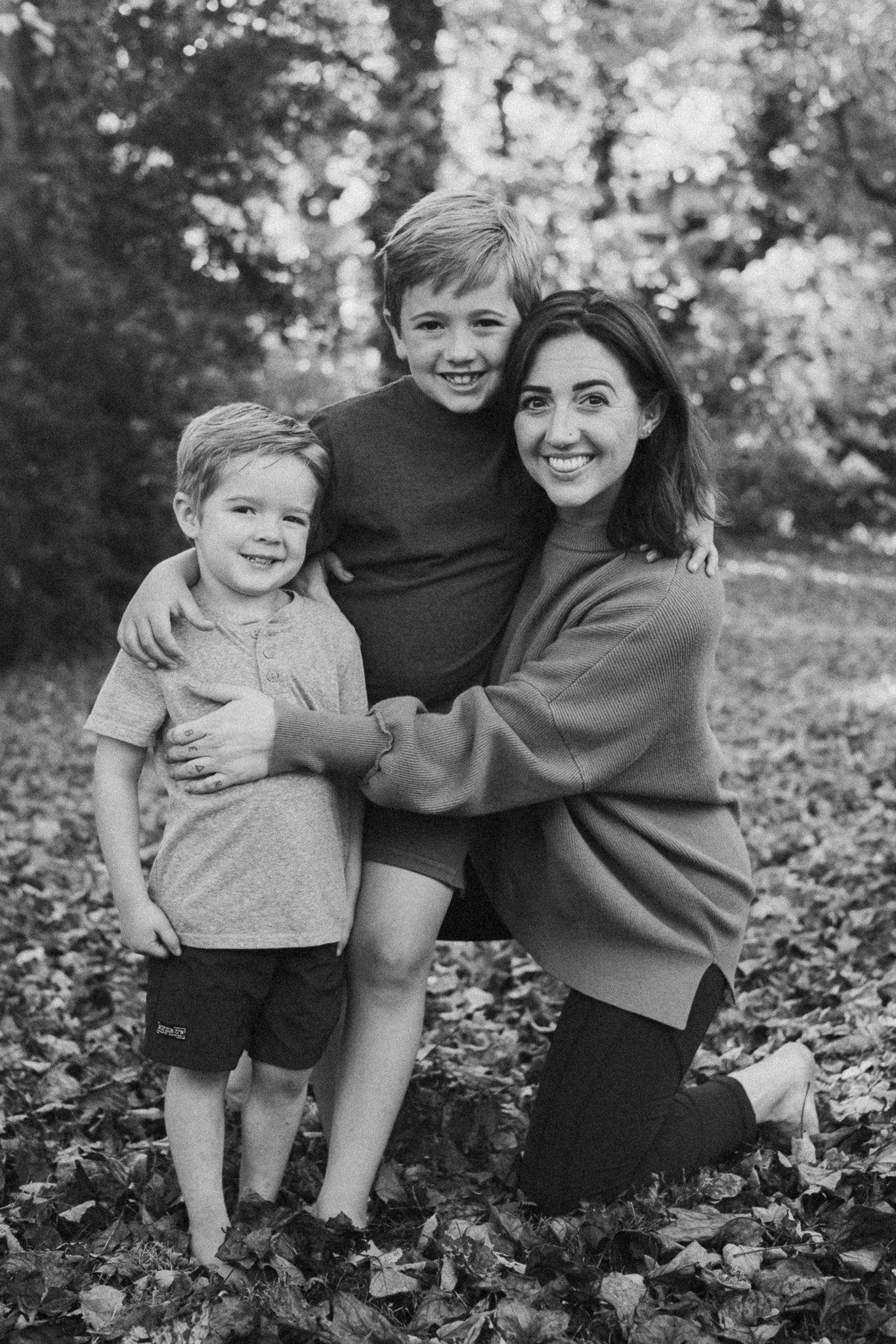 A woman and two young boys smiling outdoors among fallen leaves and trees.