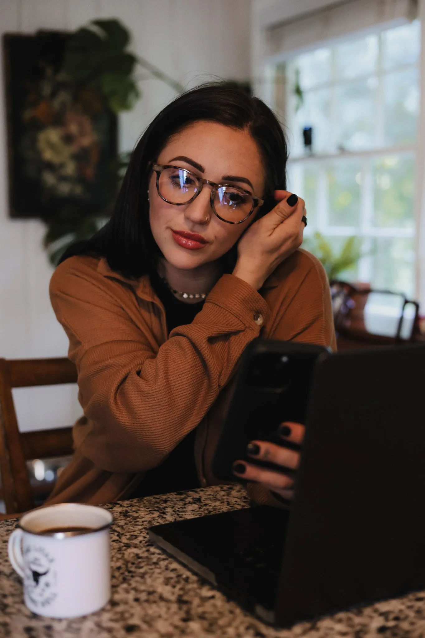 A young woman with black hair, glasses, and a nose ring sitting at a kitchen counter, looking at her phone, with a coffee mug and laptop in front of her, in a cozy, well-lit room.