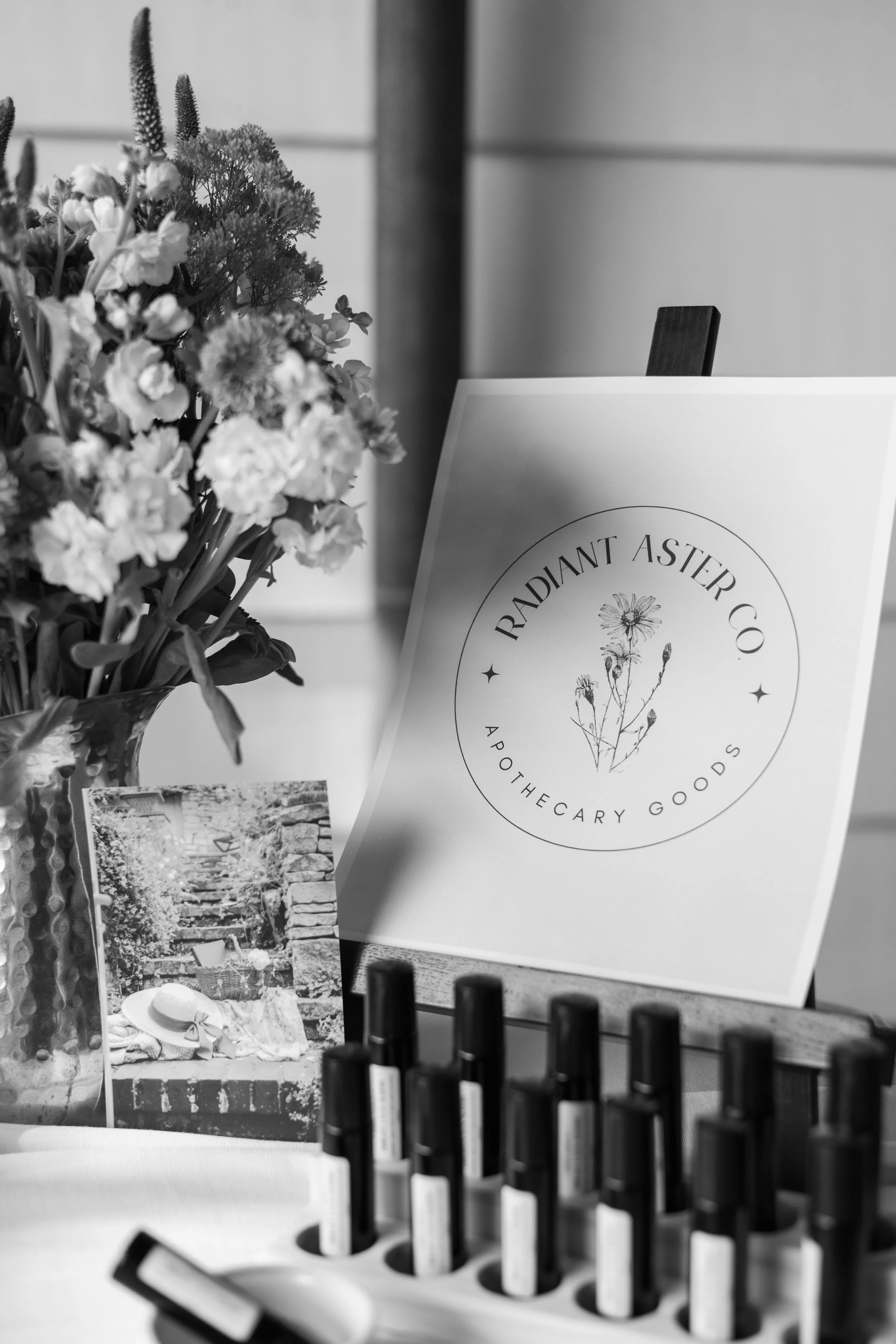 A display of aromatic essential oils in black bottles, a printed sign reading 'Radiant Aster Co., Aromatherapy Goods,' a vase of mixed flowers, and a small photo on a table.