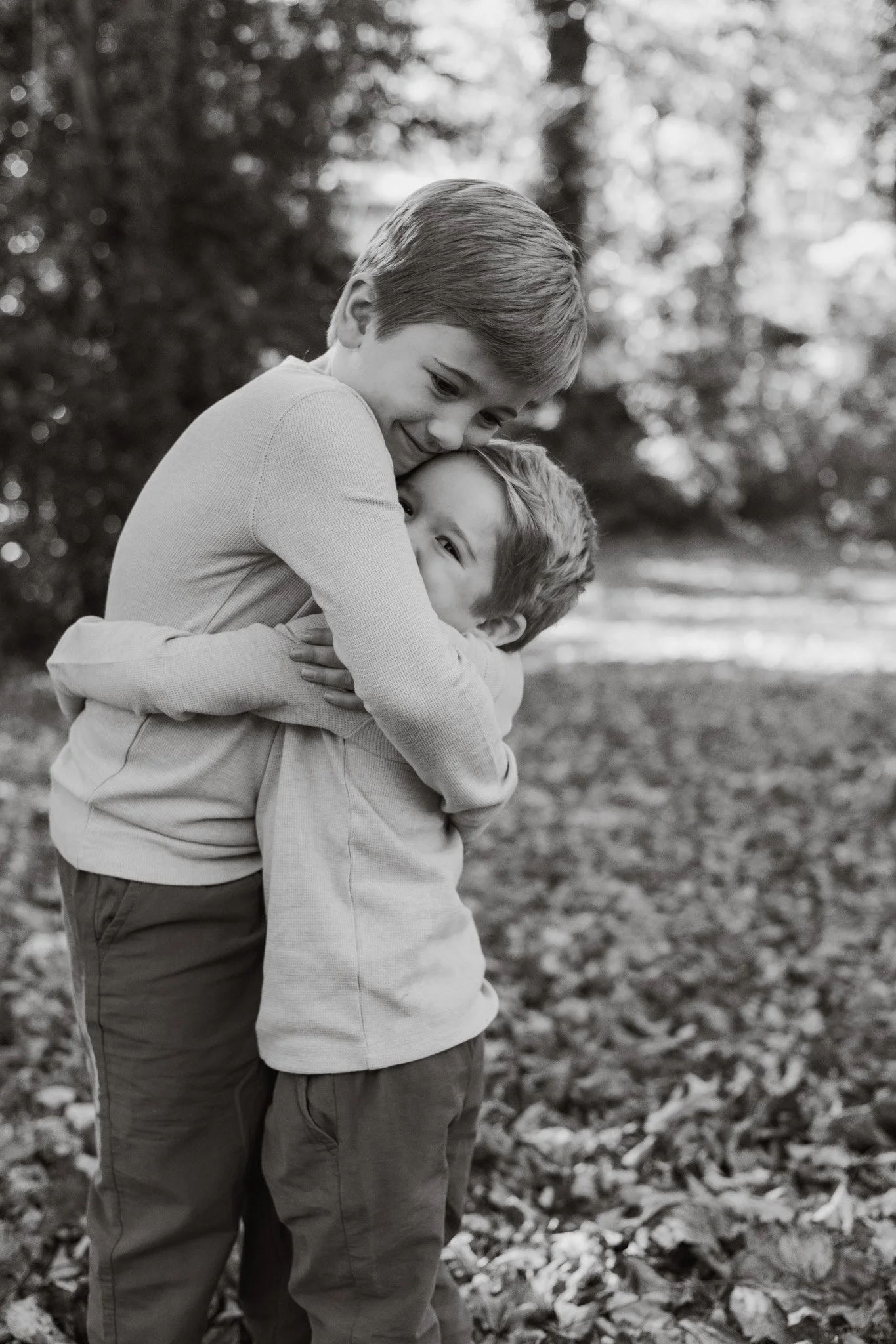 Two young boys sharing a hug outdoors on a fall day, surrounded by trees and leaves.