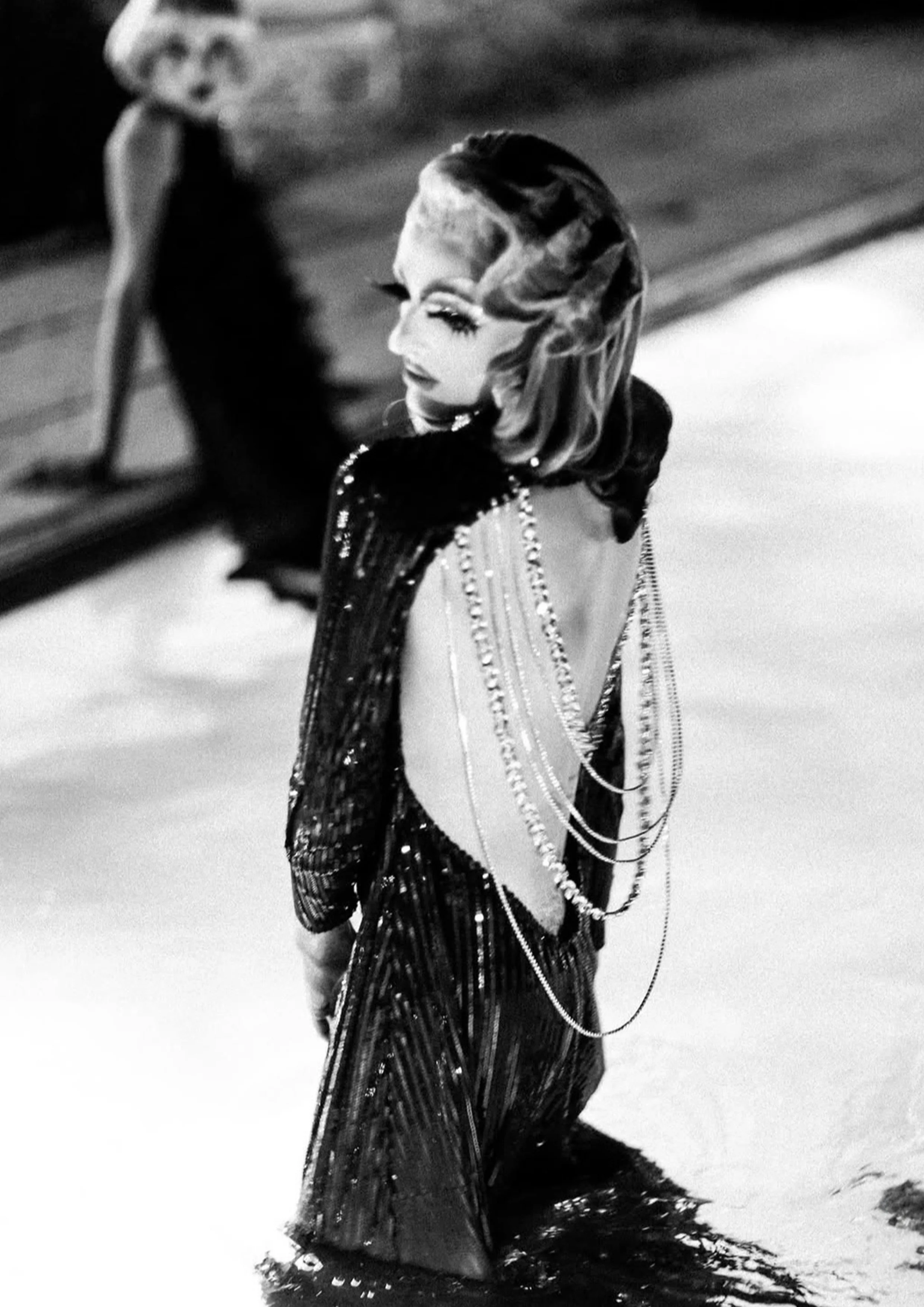 Photo artistique en noir et blanc de l'artiste drag Gala de La Nuit Drag, vue de dos, mettant en valeur une robe de soirée à paillettes dos-nu ornée de chaînes de perles.
