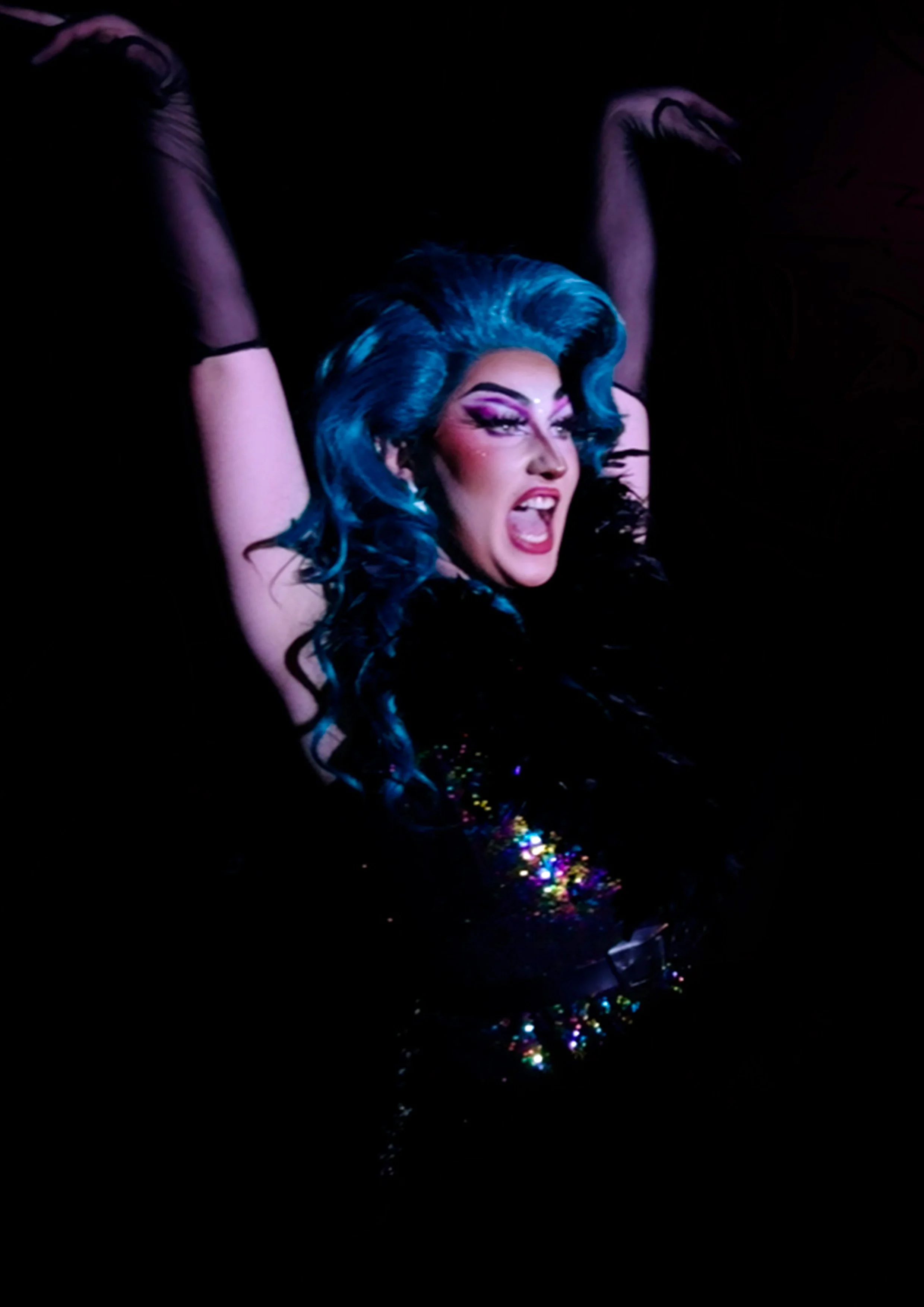 Portrait de l'artiste drag queen Kitsch de la troupe La Nuit Drag, performant sur une scène sombre avec des cheveux bleus et une robe à paillettes.