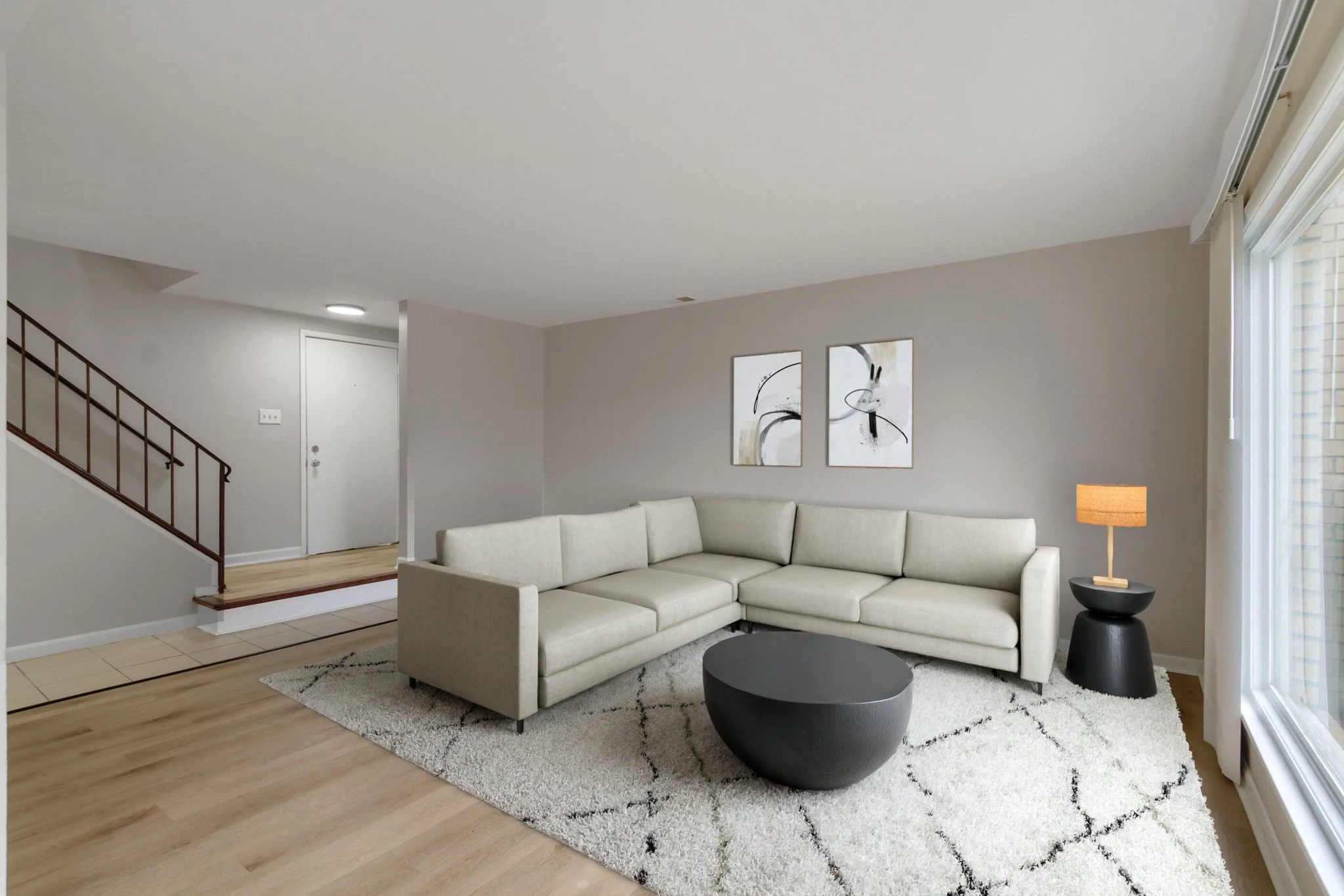 Living room with cream-colored sectional sofa, black round coffee table, black side table with a lamp, white rug with black lines, artwork on the gray wall, large window with curtains, and a staircase with wooden railing in the background.
