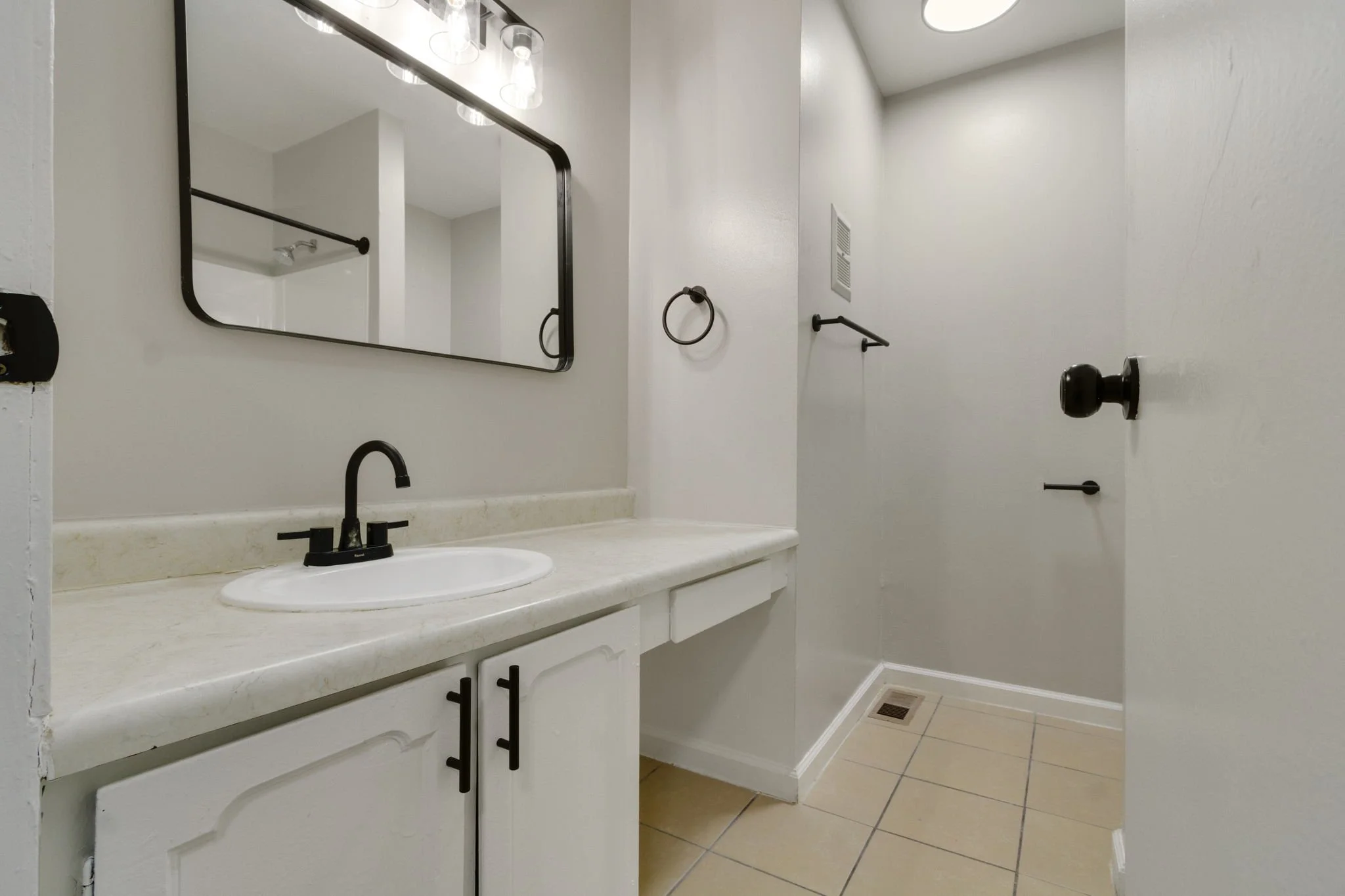 A small bathroom with white walls, a white countertop with a built-in sink, black faucet, black cabinet handles, a rectangular mirror with black frame, towel ring, towel bar, and a door with black hardware. The floor has beige tiles, and there's a ve