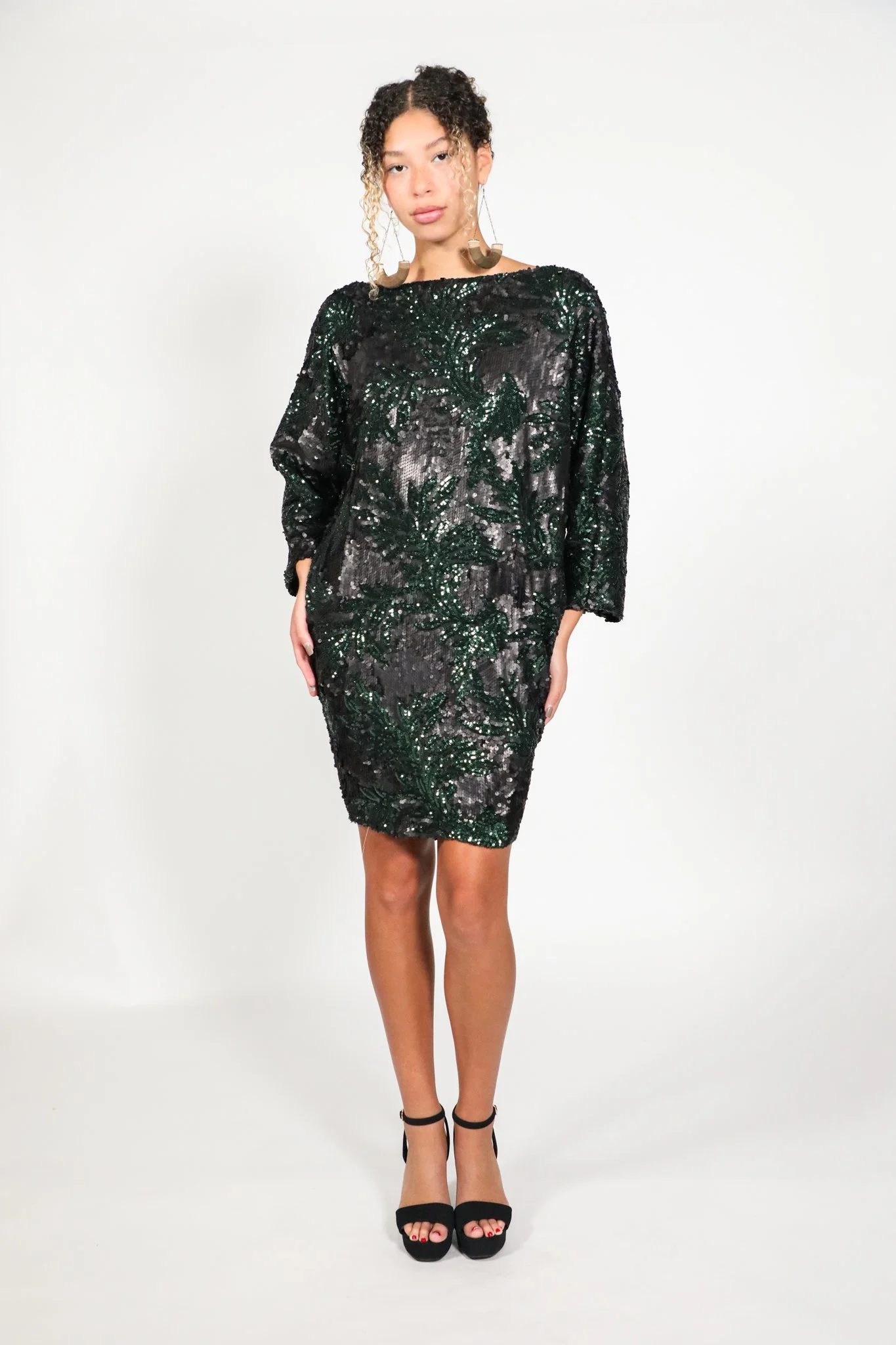 Woman wearing a black sequined dress with green leafy patterns and black high heels, standing against a plain white background.