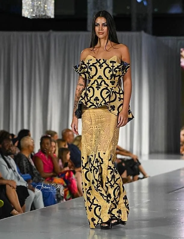 A female model walking down the runway wearing a yellow and black patterned off-shoulder top and matching high-waisted pants during a fashion show.