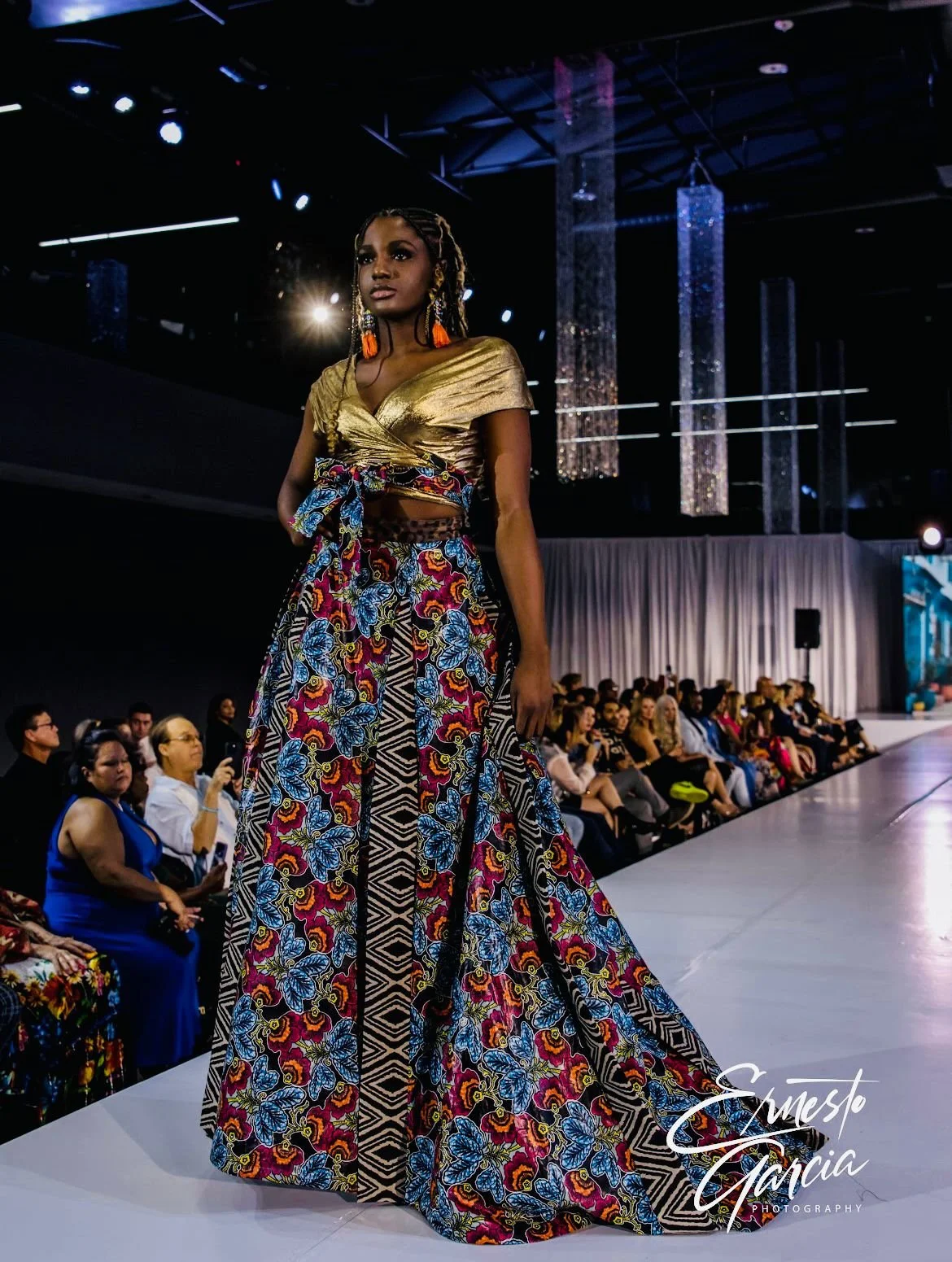 A woman walking the runway at a fashion show, wearing a gold top with a knot and long colorful patterned skirt, surrounded by seated audience members and stage lighting.