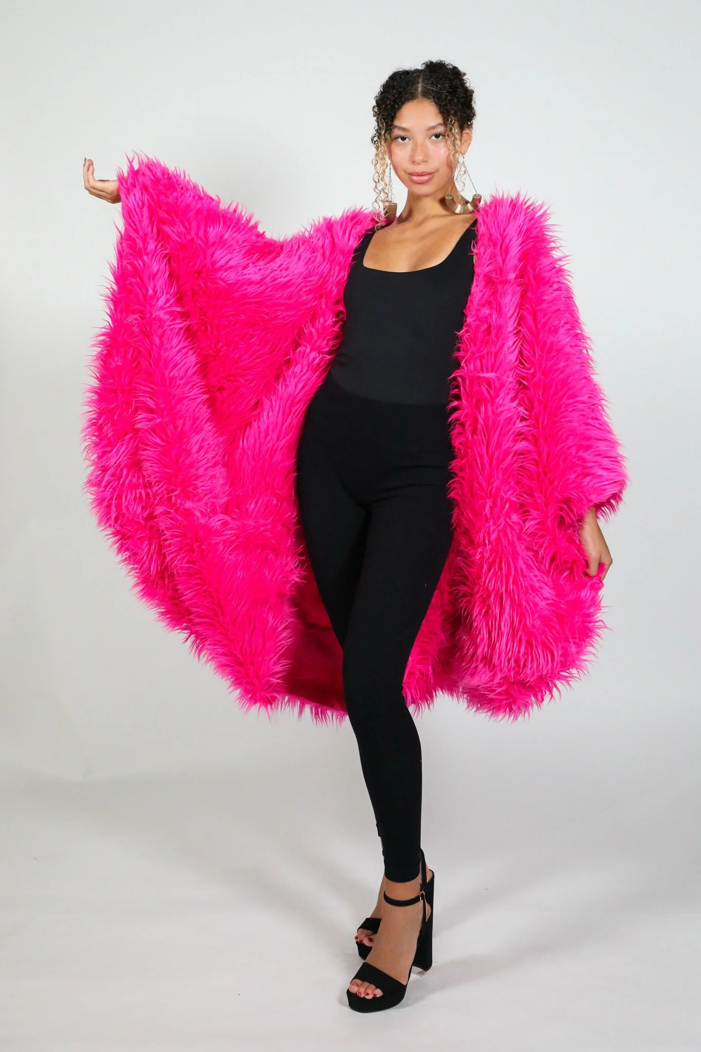 Woman wearing a bright pink furry coat, black top, black pants, and black high heels against a plain background.