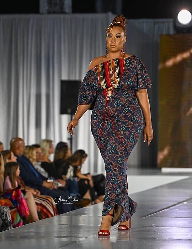 A woman walking runway in colorful patterned dress and red heels at a fashion show.